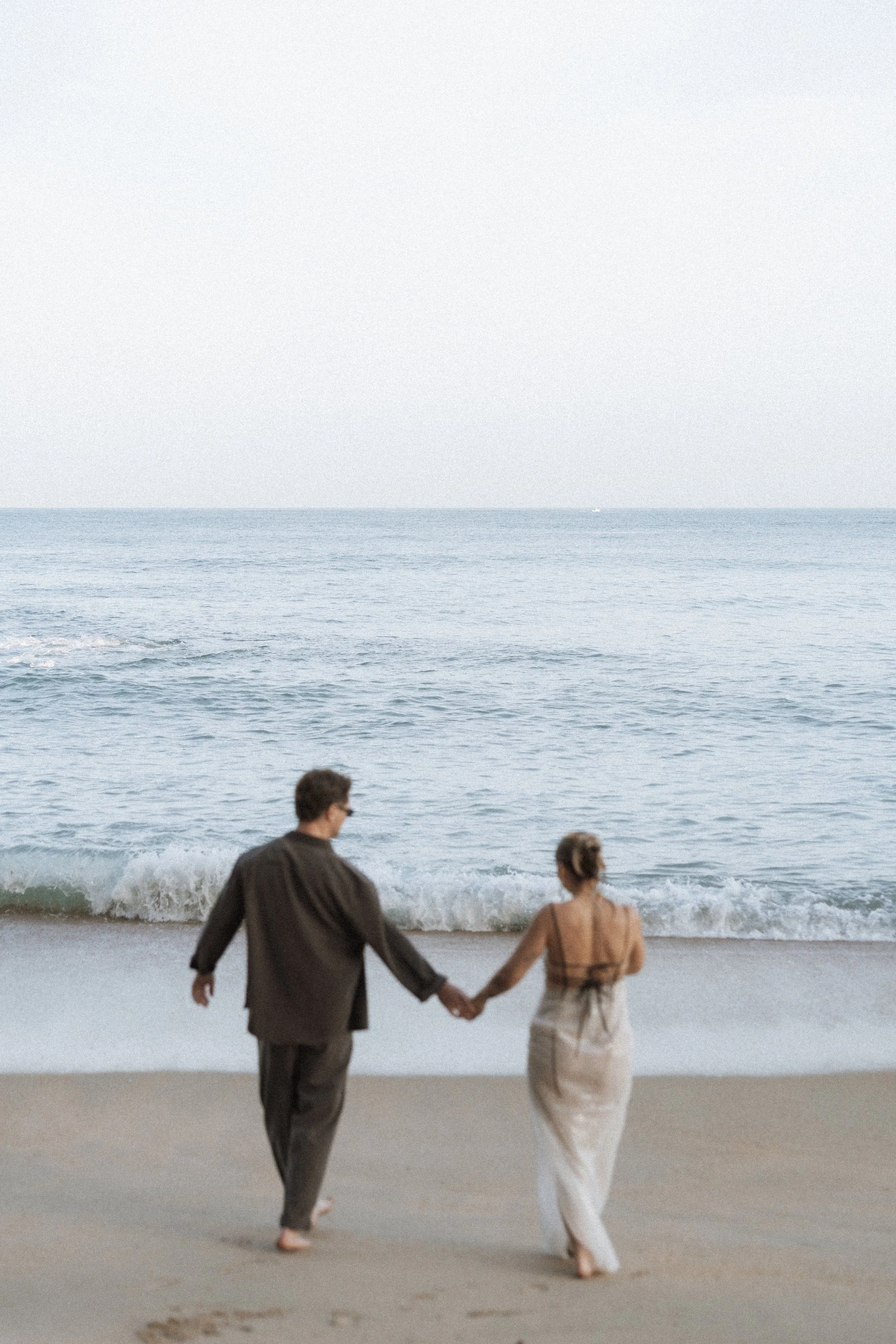 Pre-wedding couple holding hands walking towards the ocean in Biarritz, soft focus editorial beach photo with relaxed, romantic atmosphere