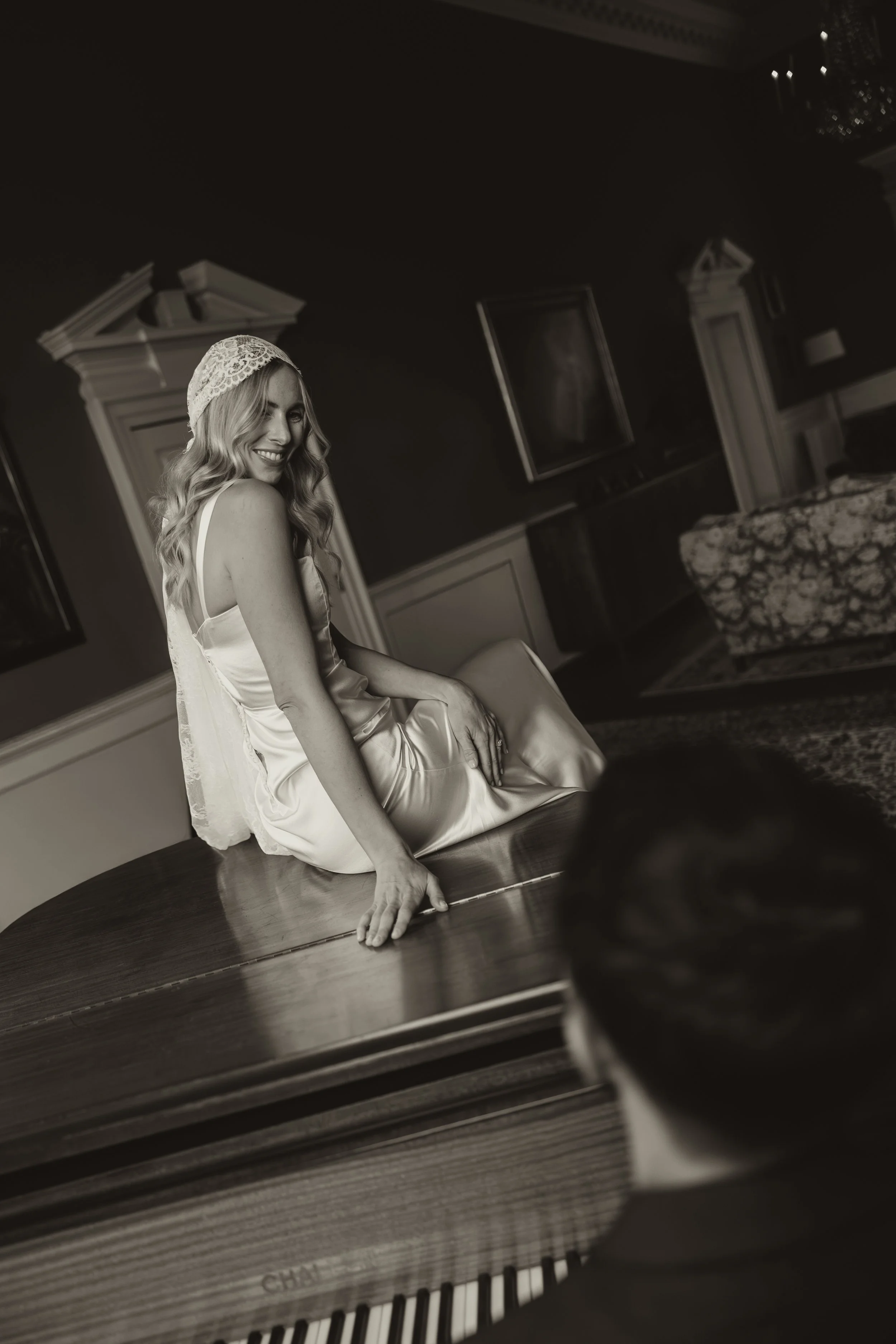 A woman in a satin slip and lace cap sitting on a table, smiling, with a man in glasses playing piano in the foreground.
