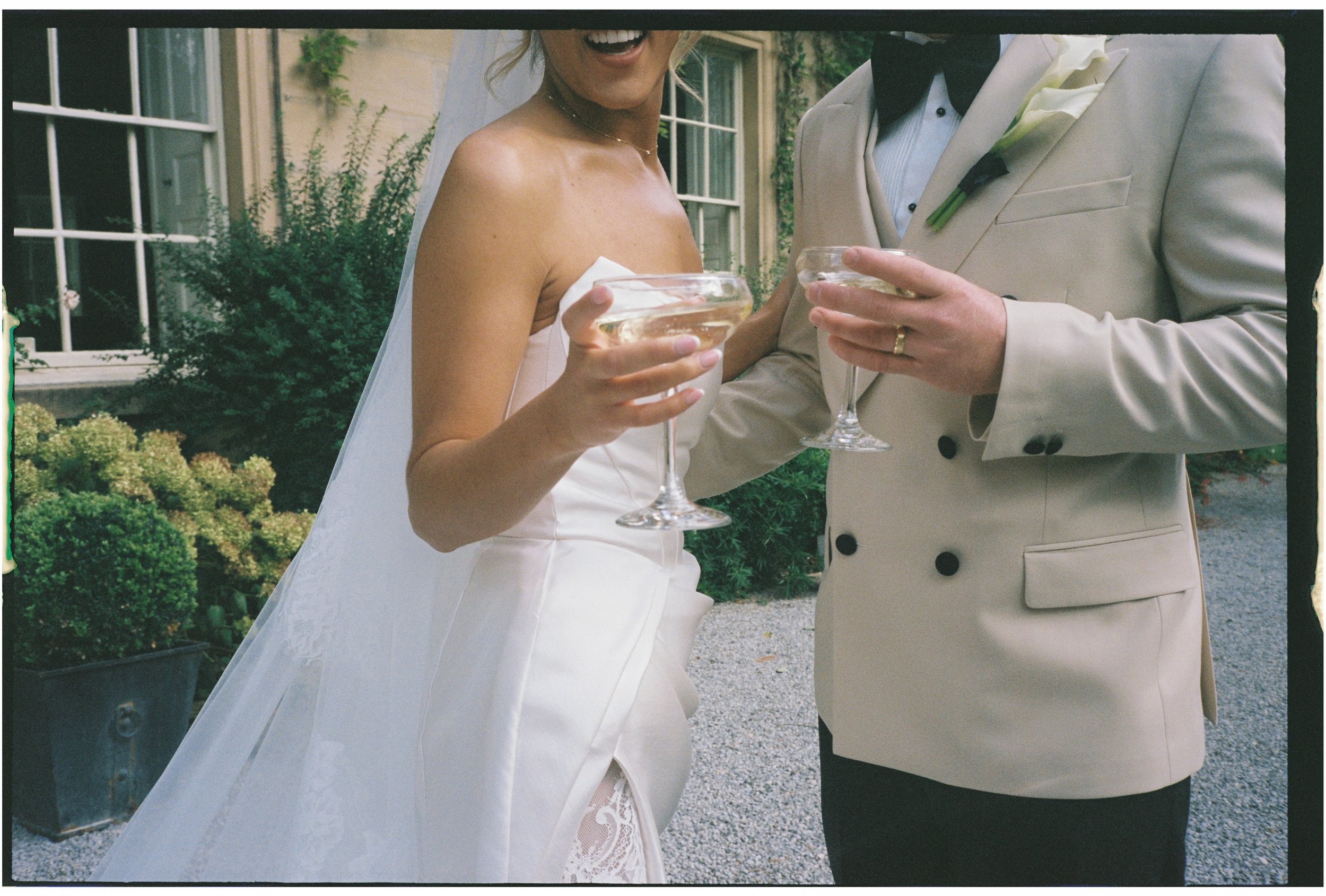 A bride and groom in wedding attire holding champagne glasses during a celebration outside a house with greenery.