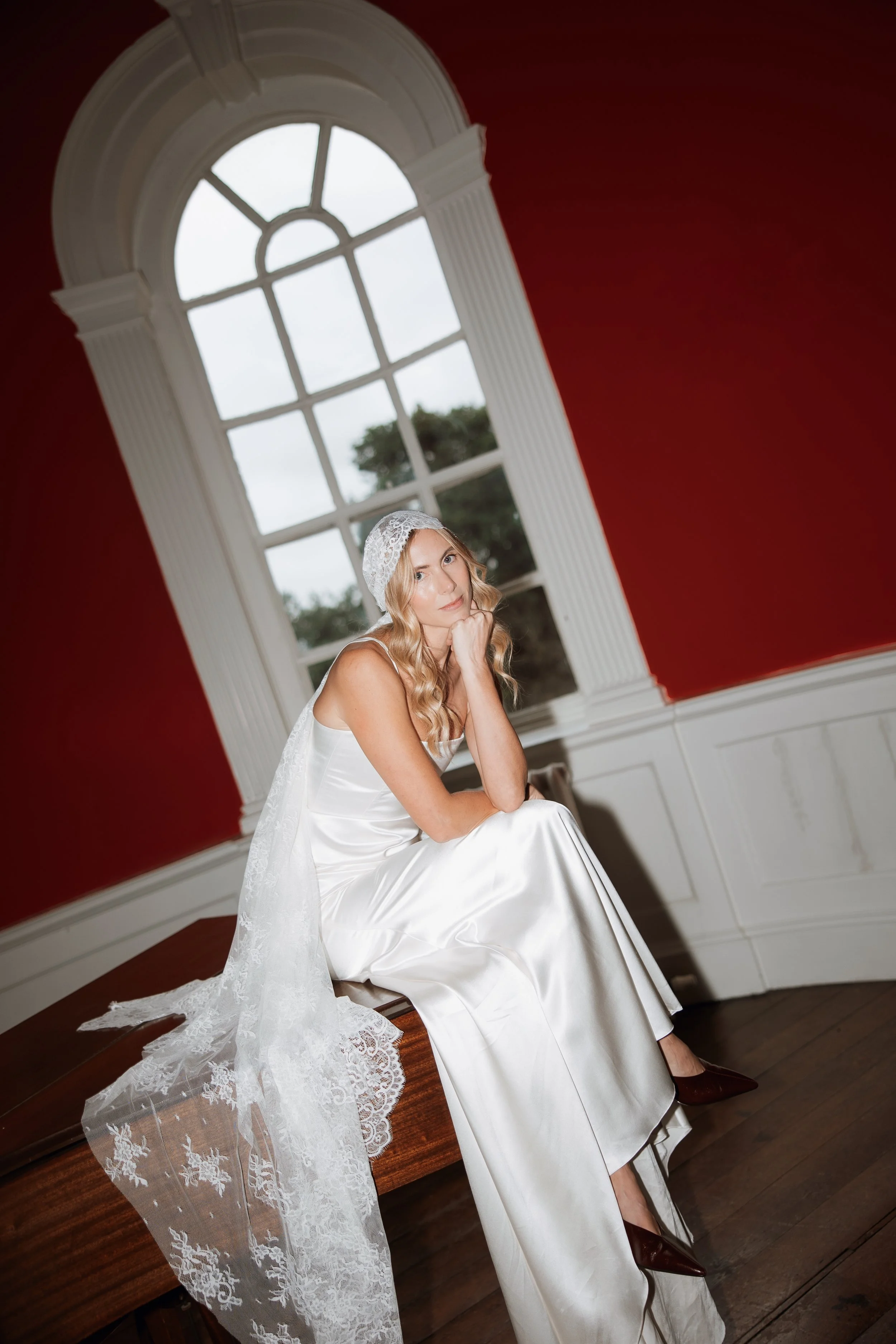 A woman in a white silk wedding dress with a lace veil, sitting on a piano indoors by a large arched window with white trim, red walls, and a view of trees outside. Taken at Iscoyd Park wedding venue.