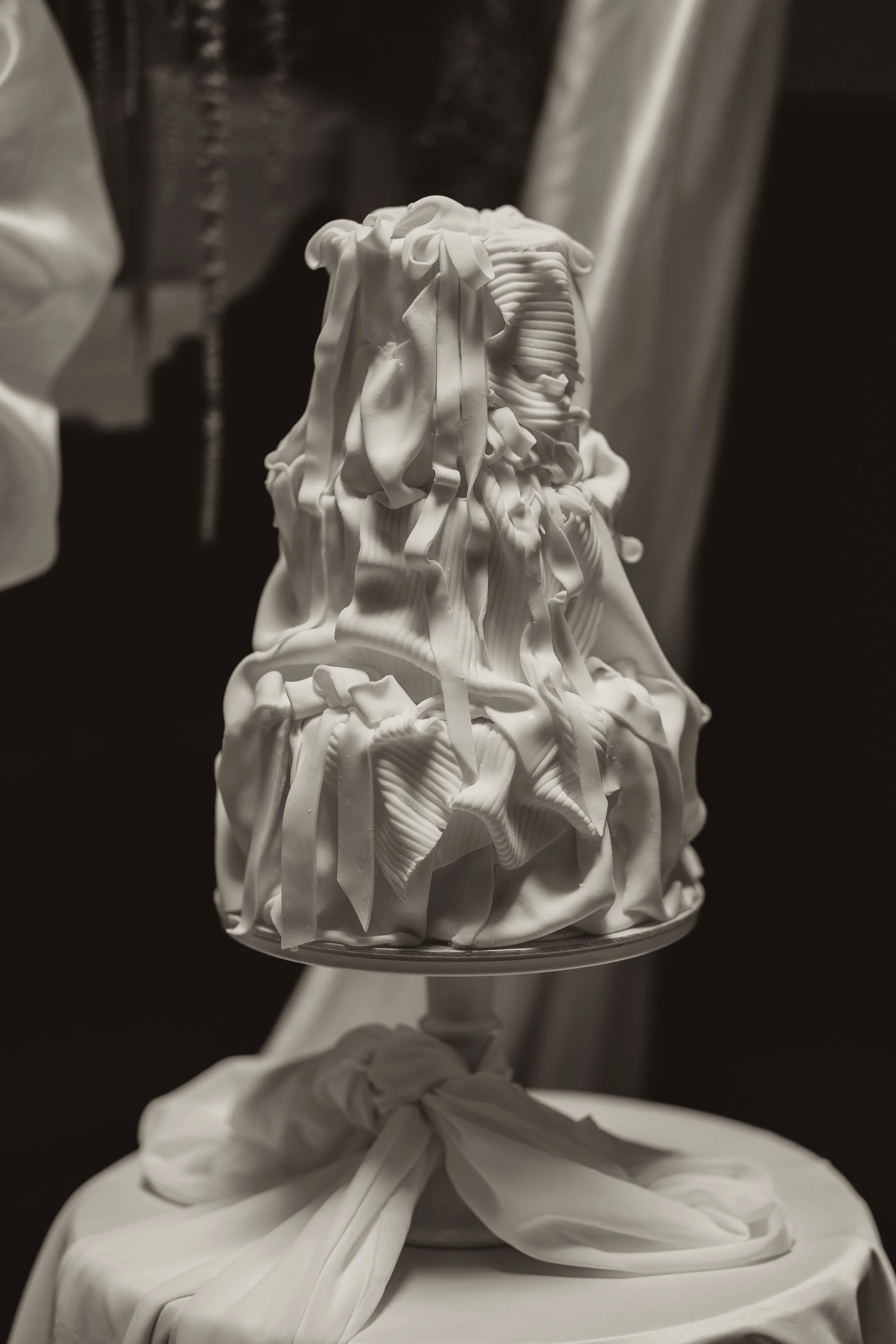 A sculptural cake decorated with intricate white fondant with ribbon-like and textured details, topped with a bow, placed on a stand with a draped white cloth