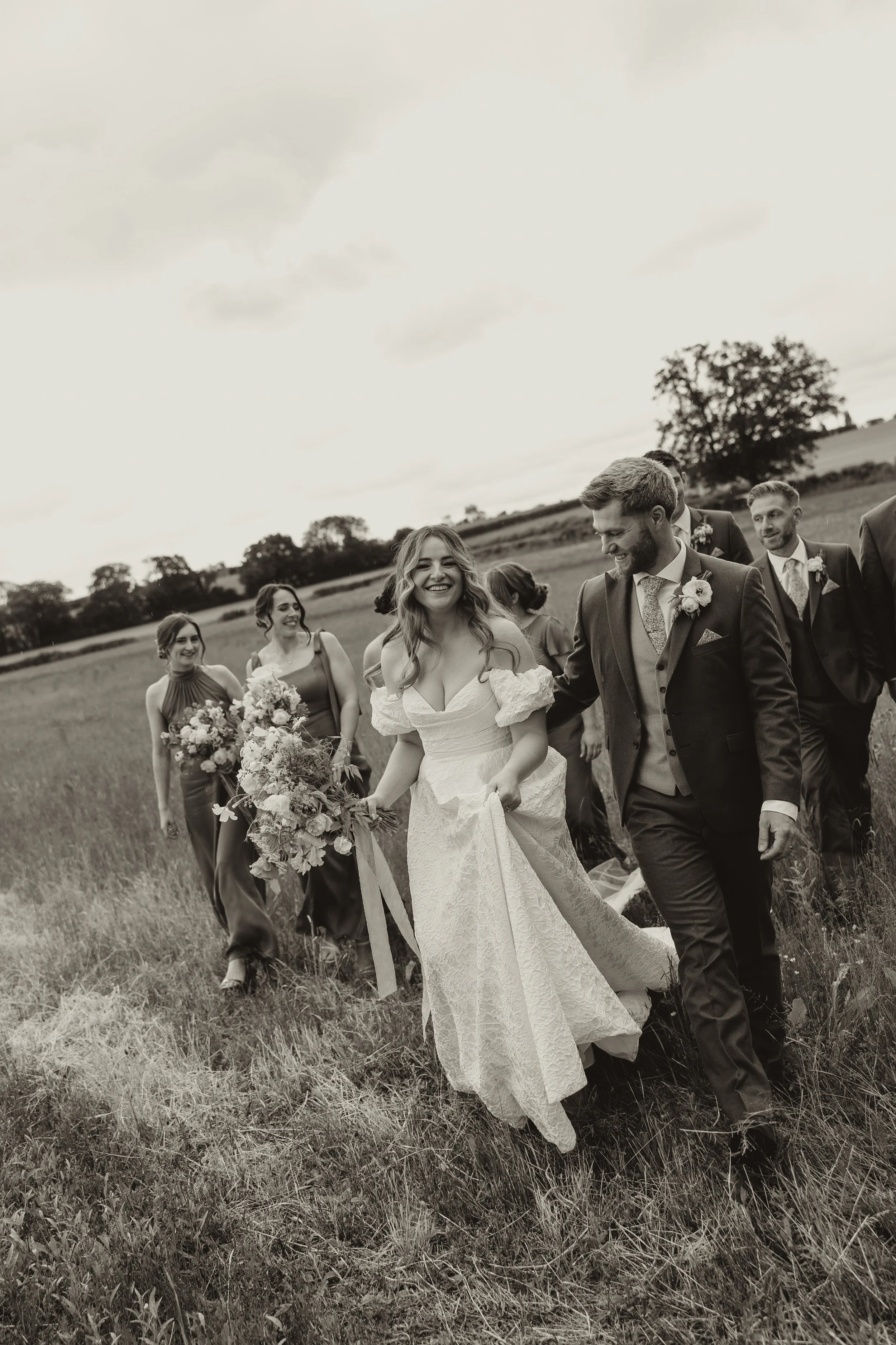 A black and white photo of a wedding party walking through a grassy field, with a smiling bride in a white lace gown, holding a bouquet, and a groom in a dark suit, surrounded by bridesmaids and groomsmen in formal attire.