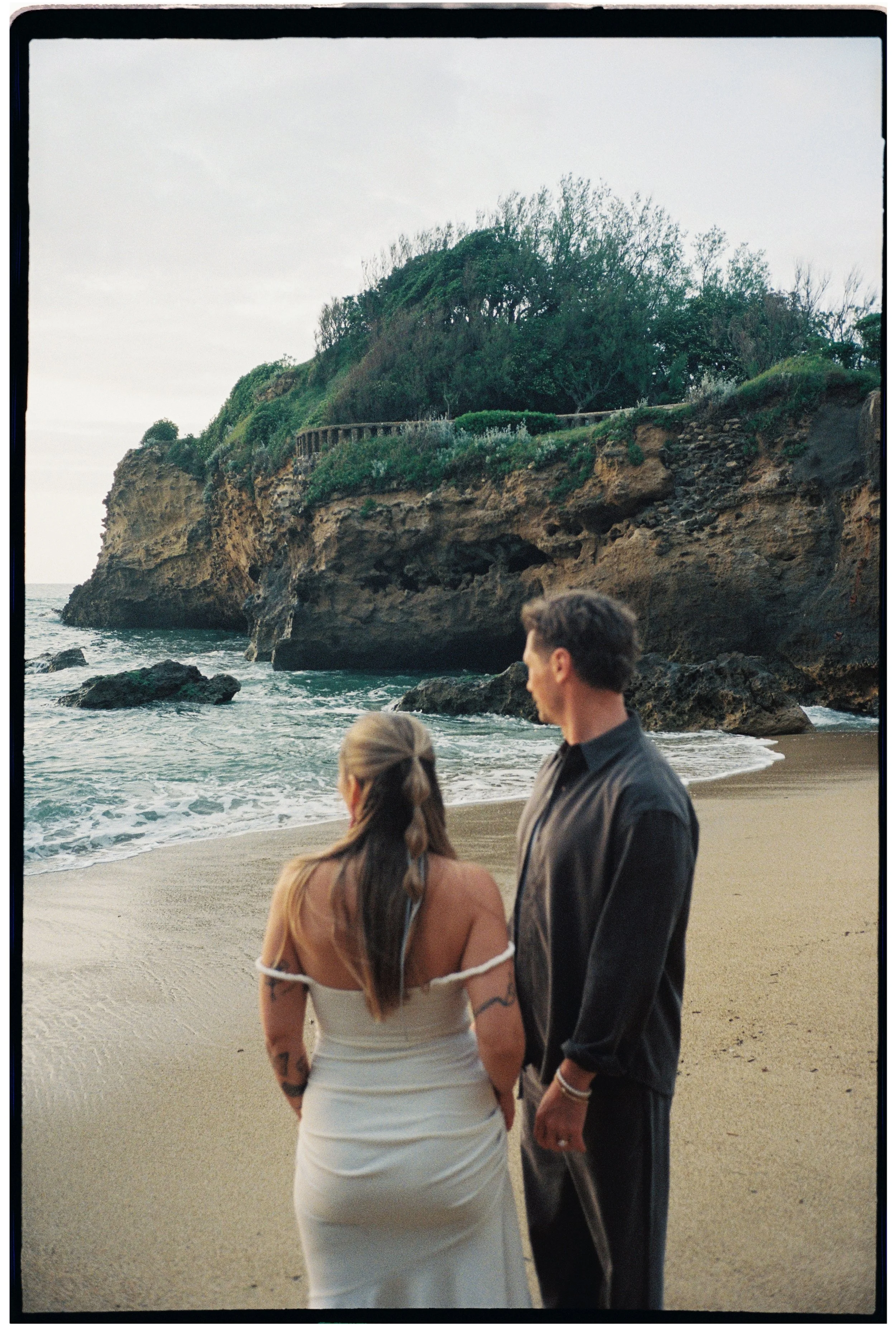 Couple standing together on a quiet beach at sunset with dramatic cliffs in the background, photographed on film.
