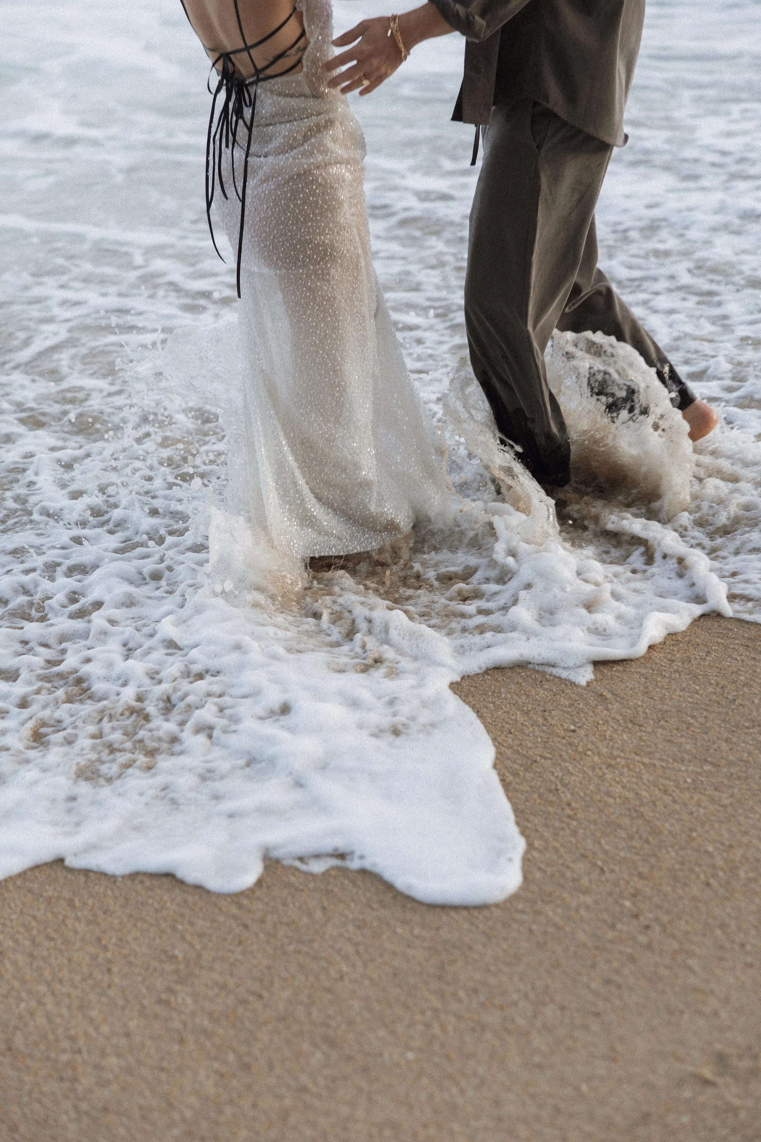 Editorial wedding photograph of couple walking through the sea on a beach, capturing a modern, fashion-inspired destination wedding moment