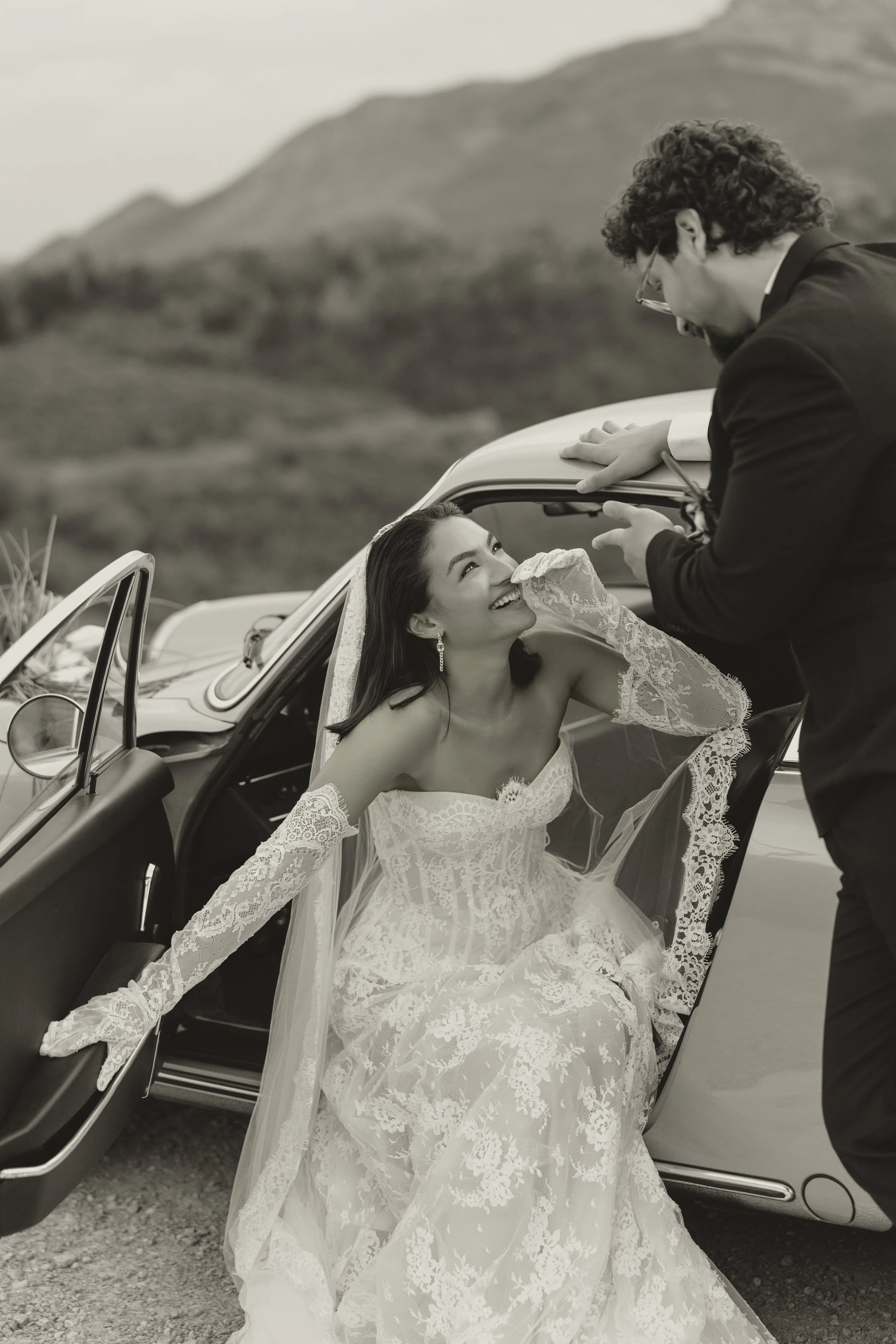 Luxury wedding couple portrait featuring a bride in a lace wedding dress and a groom beside a vintage car, photographed in an elegant editorial style