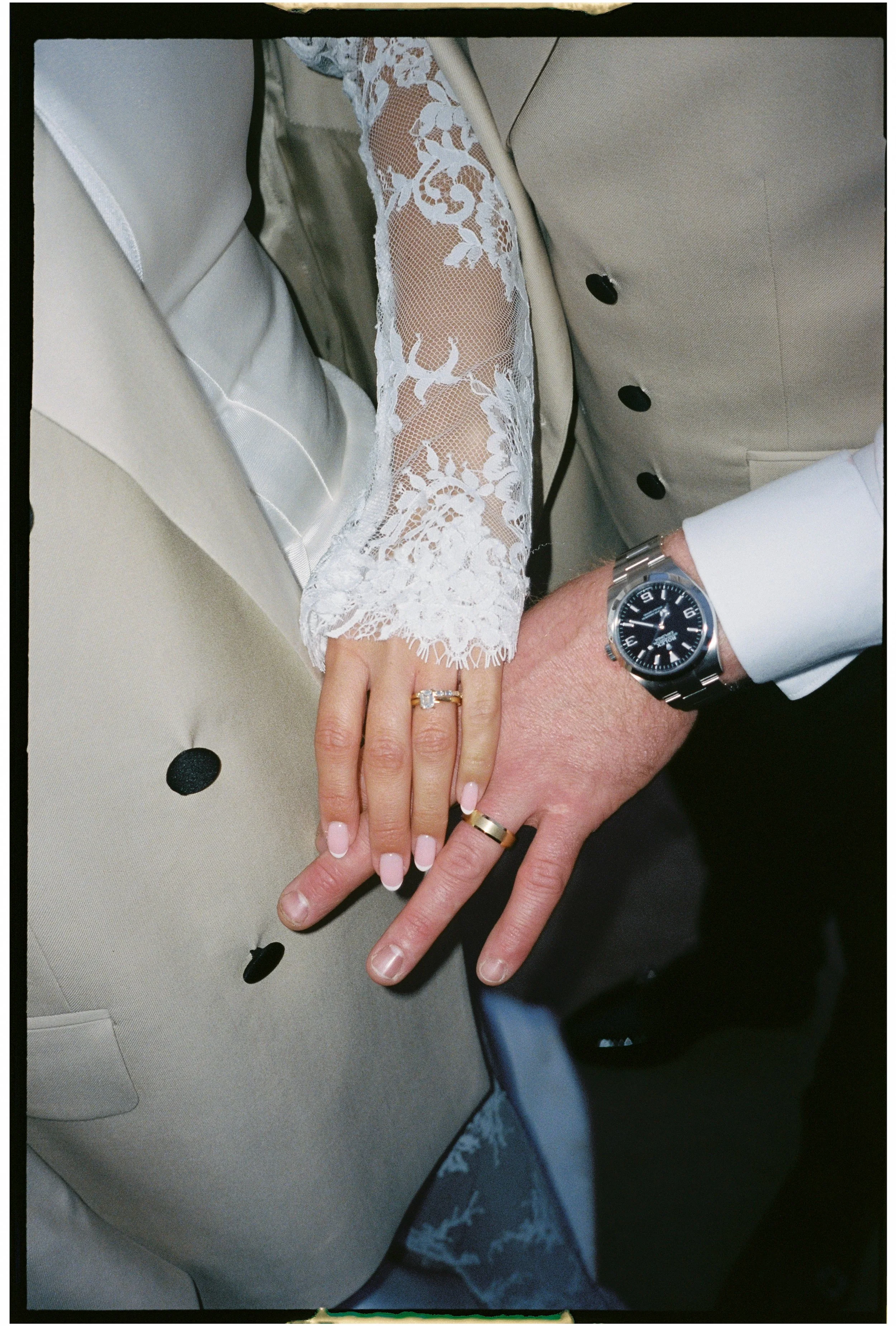 Close-up of a couple's hands showing wedding rings, with the woman's hand on top of the man's, both resting on beige suits. The woman's hand has a diamond ring and a gold band, and the man's hand has a plain gold band. The woman has manicured nails w