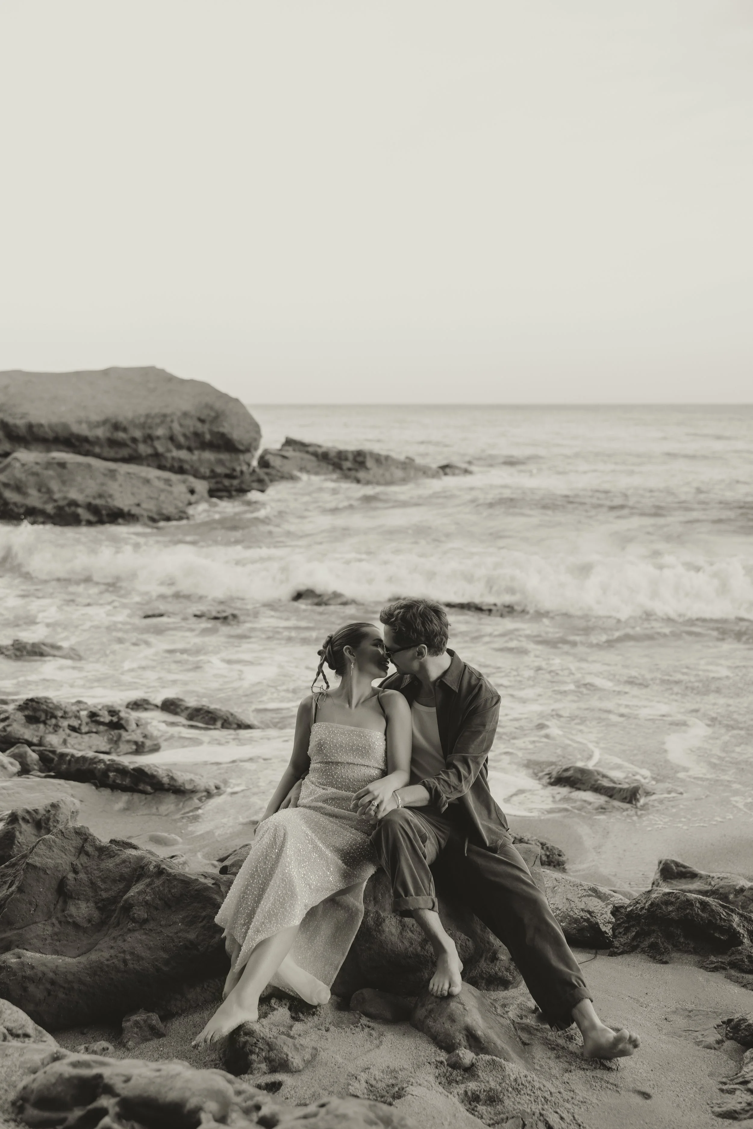 Pre-wedding couple sitting on rocks by the ocean in Biarritz, intimate black and white editorial portrait with waves in the background