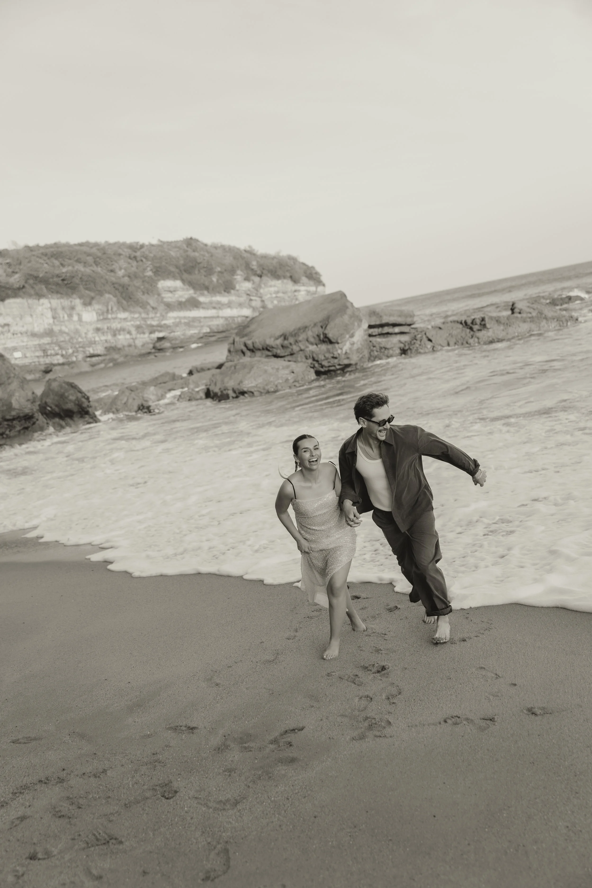 Couple running barefoot along the shoreline in Biarritz during a relaxed pre-wedding shoot, laughing together with waves washing in around them.