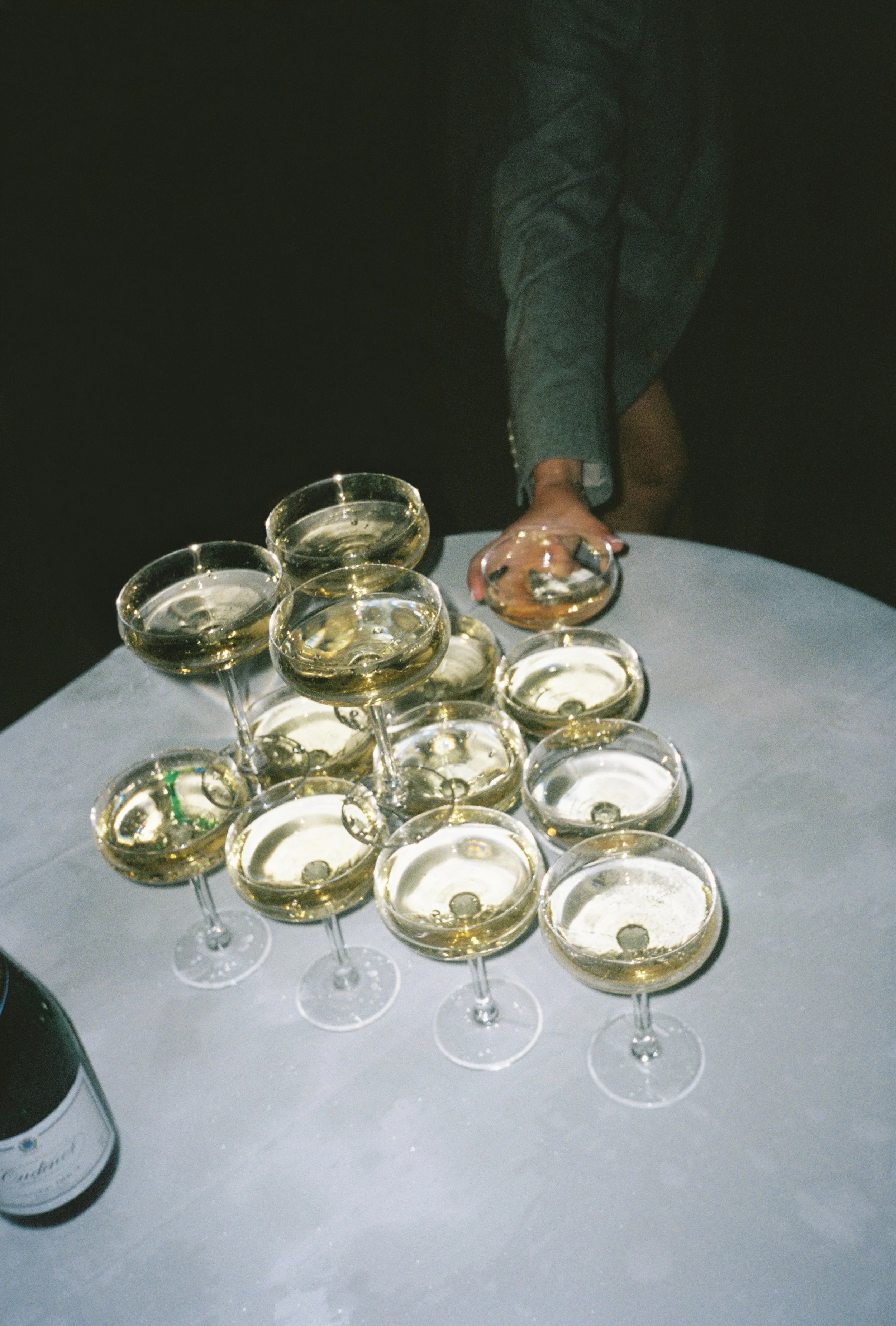 A person in a suit is reaching for a glass of champagne on a white table with multiple champagne glasses filled with champagne and a bottle of champagne on the table. Editorial 35mm film wedding photography.