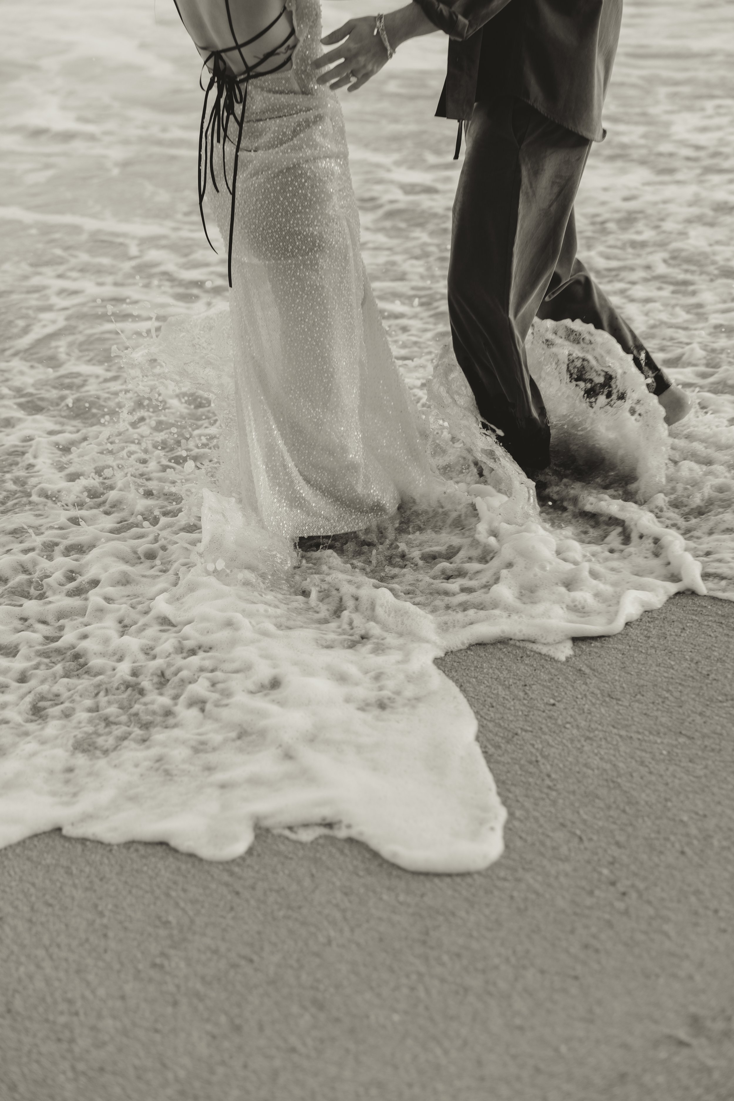 Close-up of couple walking through sea foam at the shoreline, bride’s embellished dress trailing in water during a pre-wedding shoot in Biarritz