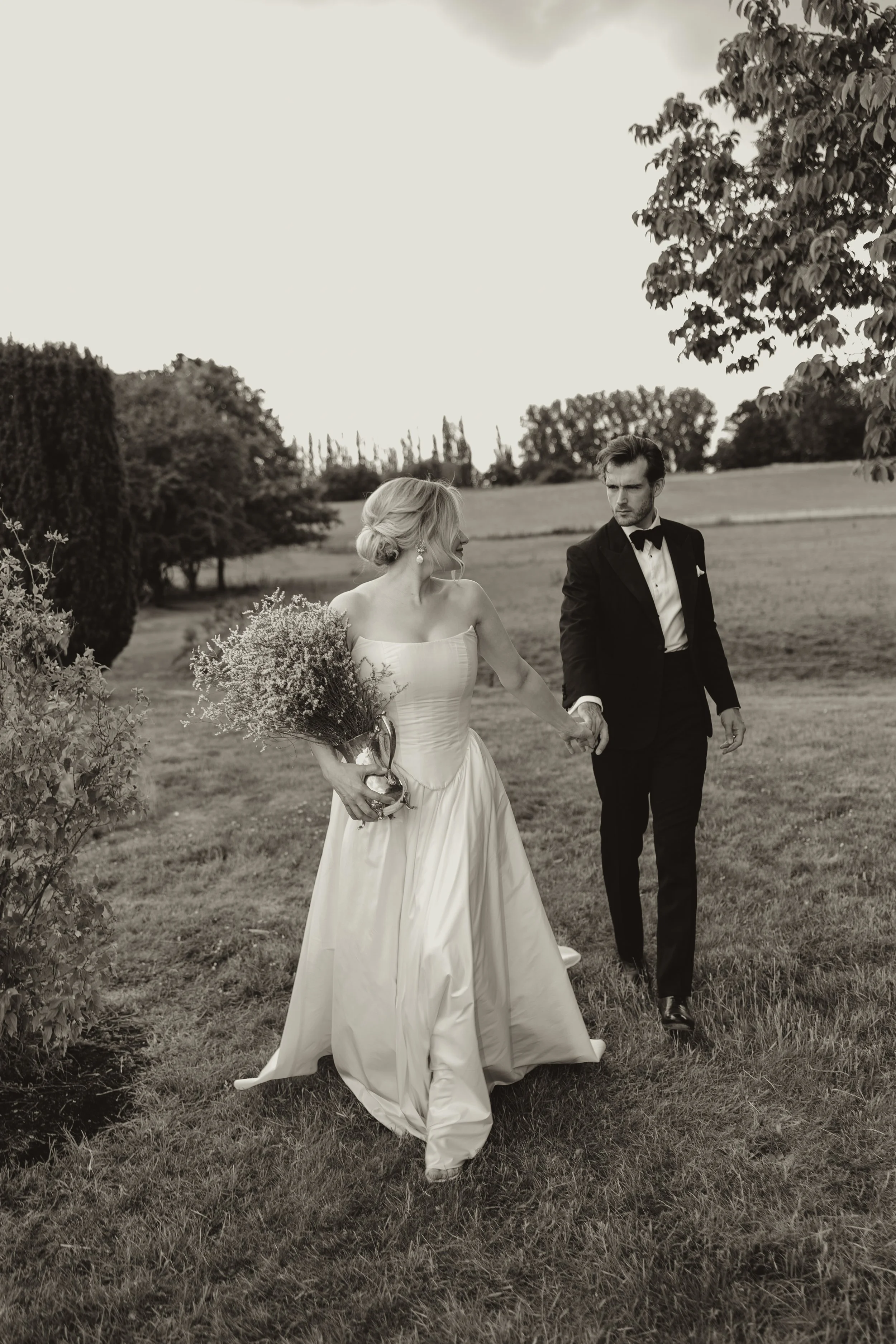 A bride in a white wedding gown holding a bouquet and a groom in a tuxedo walking hand in hand outdoors in a park or garden setting.