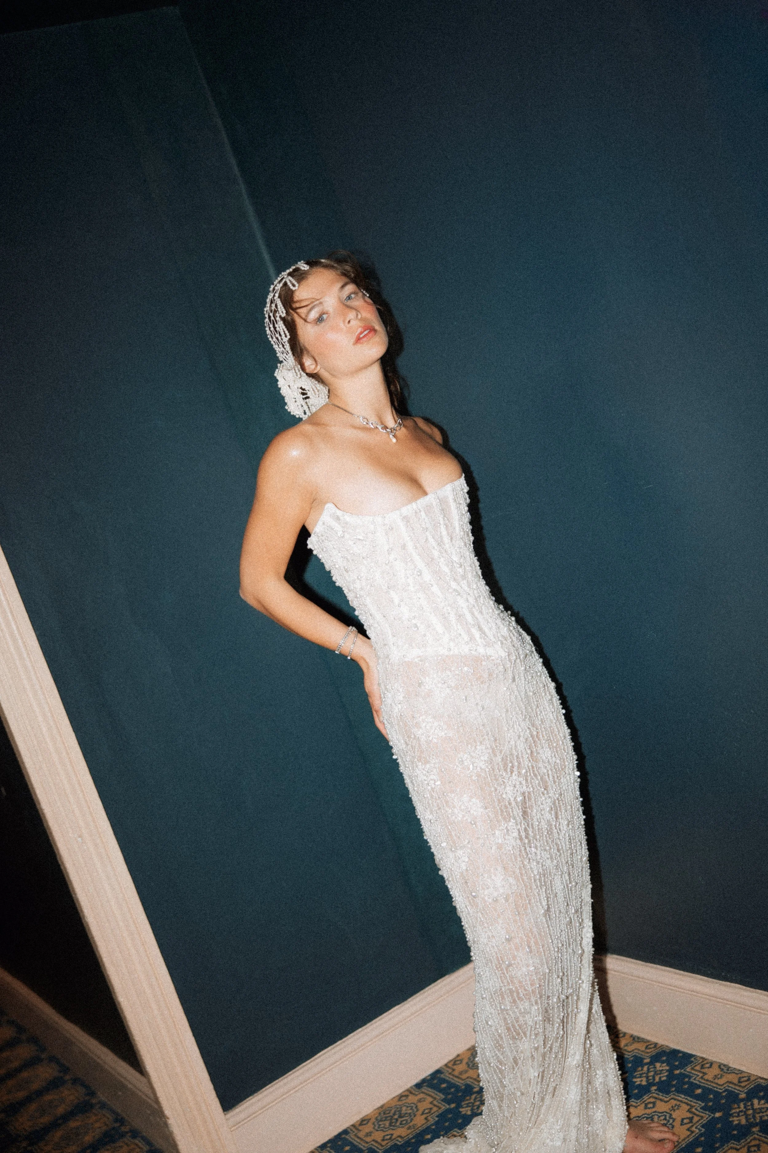 Bride in a strapless, beaded, white formal gown and jewellery poses indoors against a dark wall.