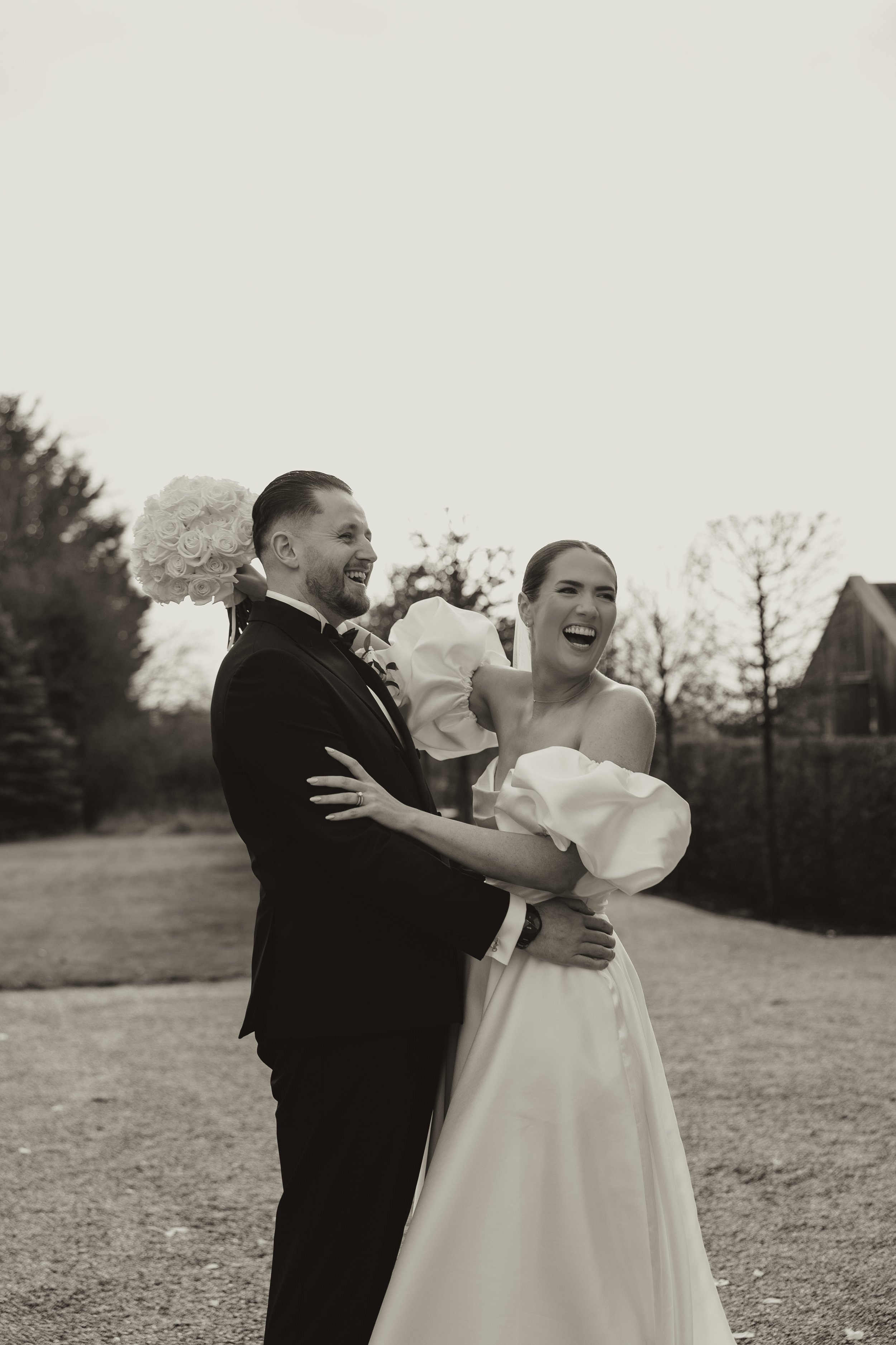 A black and white photo of a bride and groom laughing outdoors, with the groom holding a bouquet of roses on his shoulder.