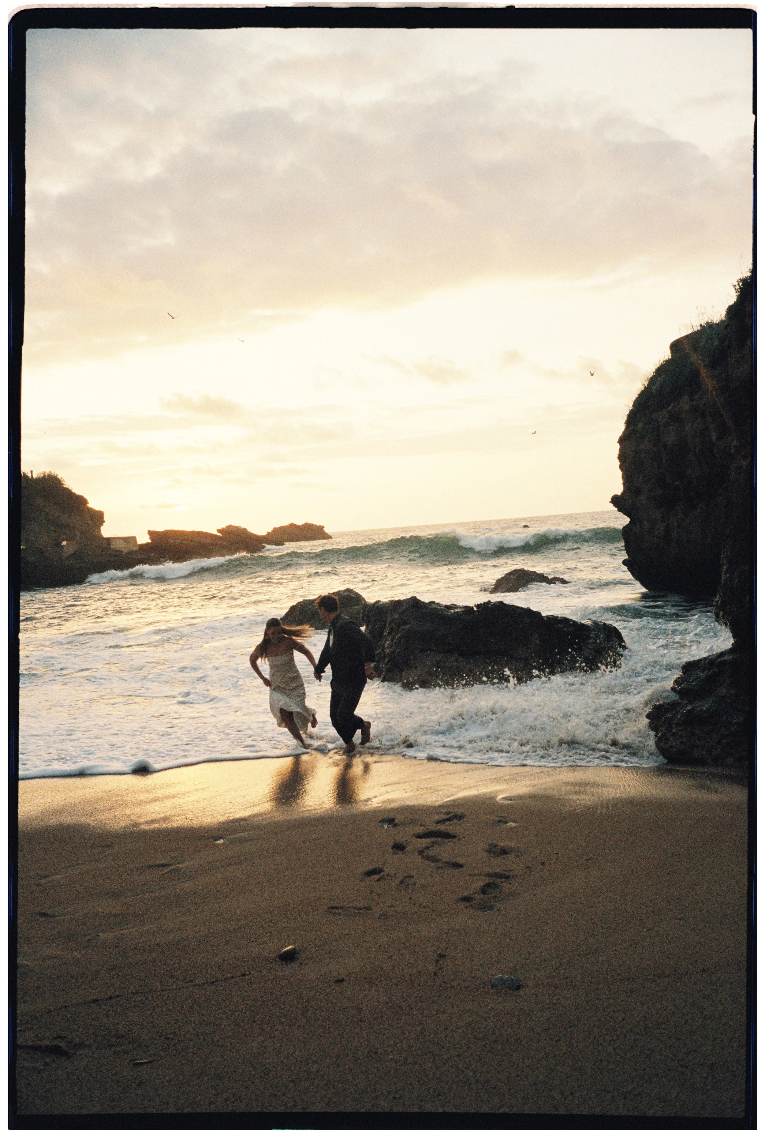 Couple running into the sea at sunset on a rocky beach during an intimate elopement session on 35mm film