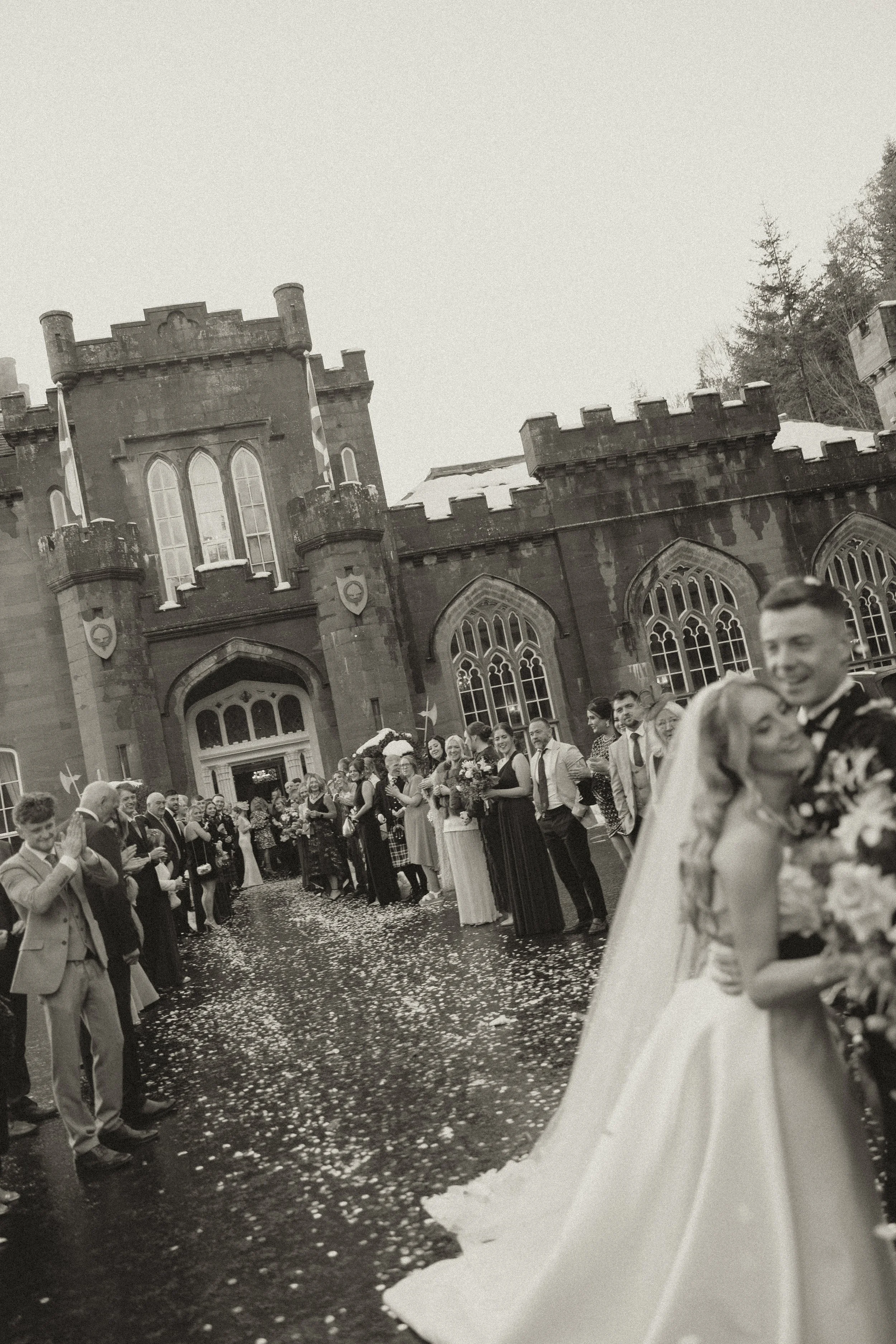 Black and white photo of a wedding outside a castle-like building, with the bride and groom in the foreground hugging, surrounded by guests lined along the pathway.