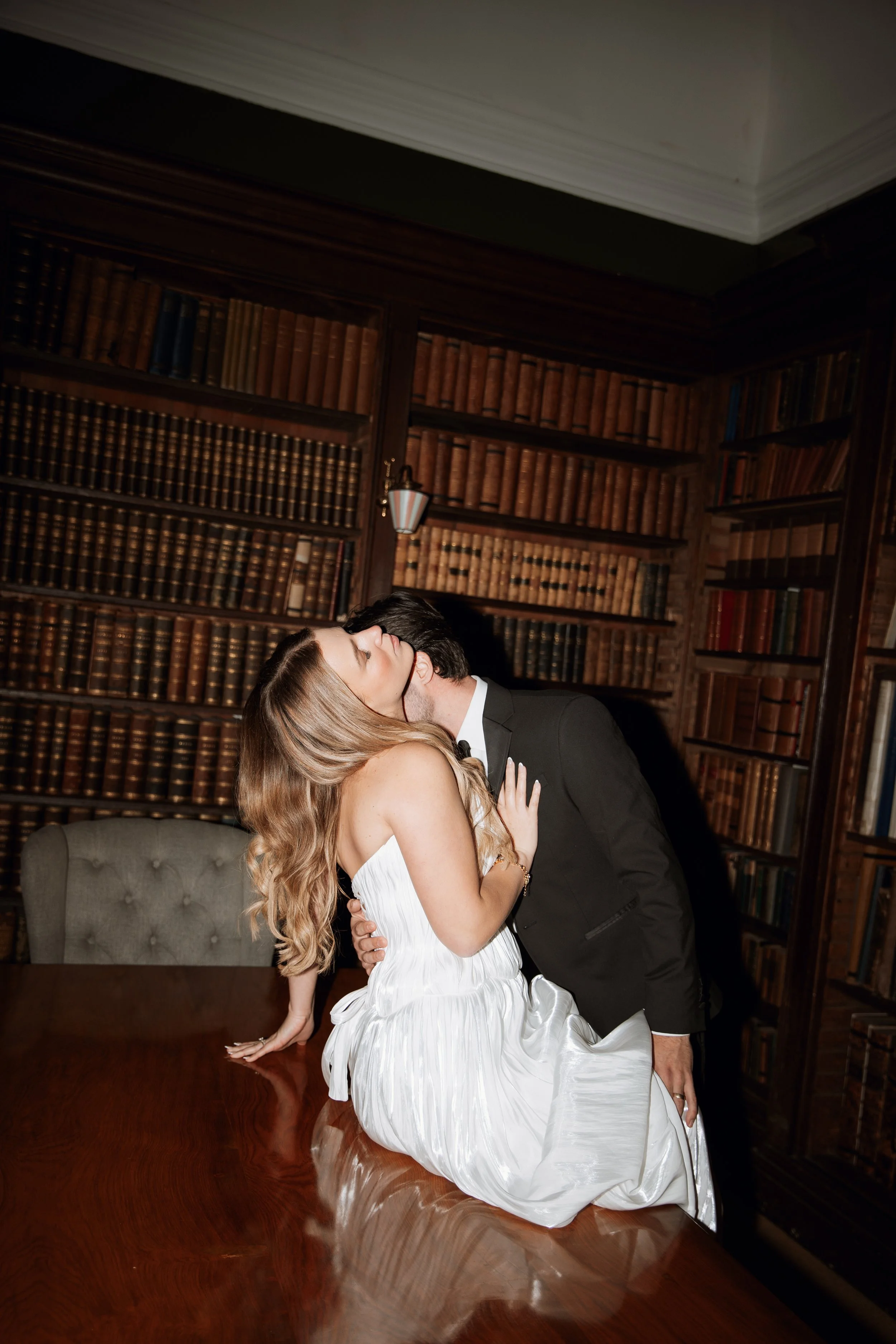 A woman in a white dress and a man in a black tuxedo kissing in a library with wooden bookshelves.