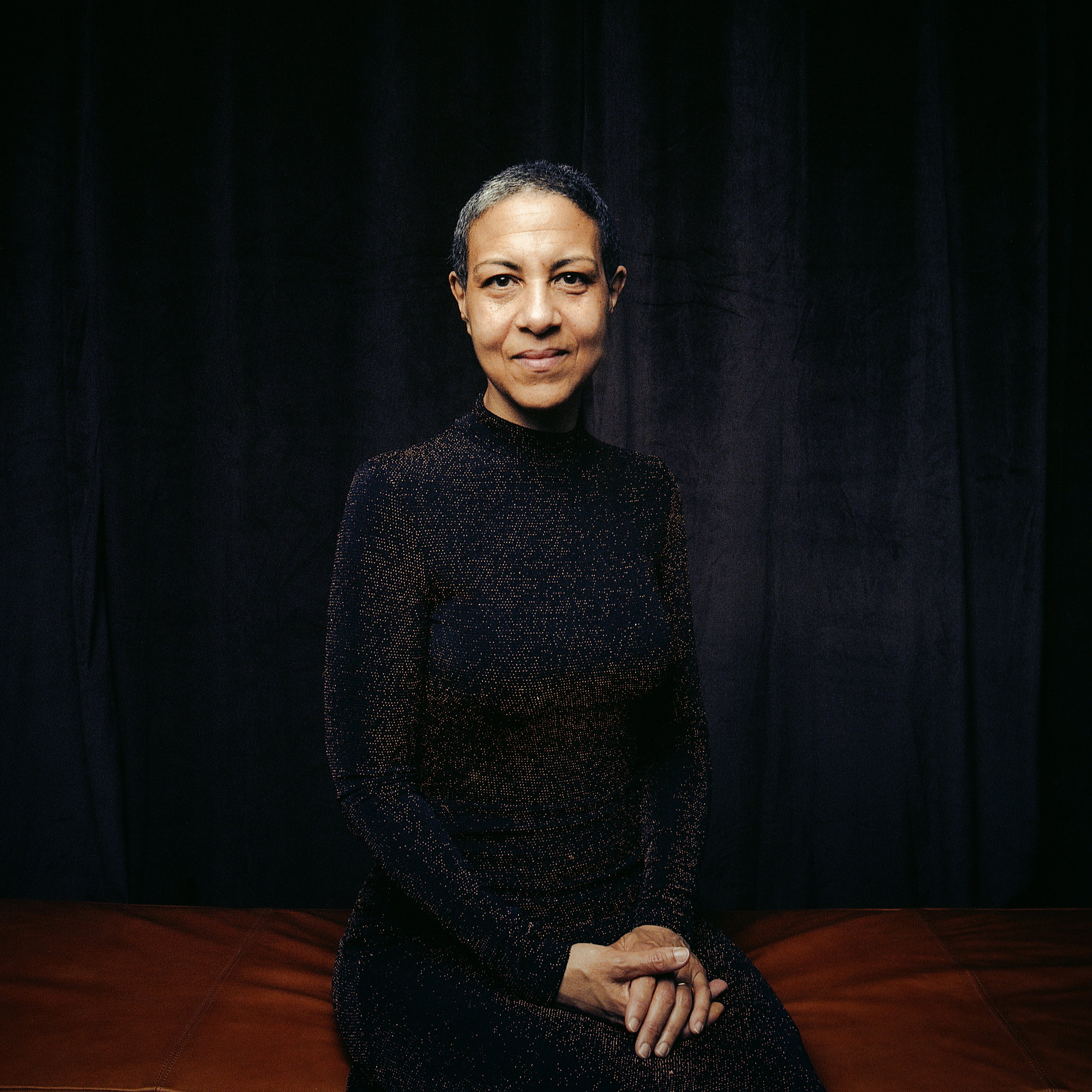 Portrait of a woman with short hair, sitting against a black curtain background, wearing a black, glittery long-sleeve top, with hands resting on her lap.