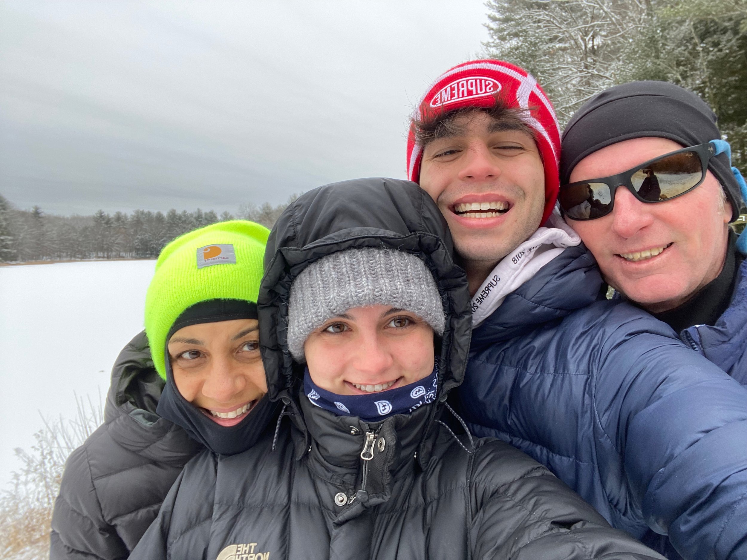 Four people bundled in winter jackets and hats, taking a group selfie outdoors in a snowy area with trees in the background.