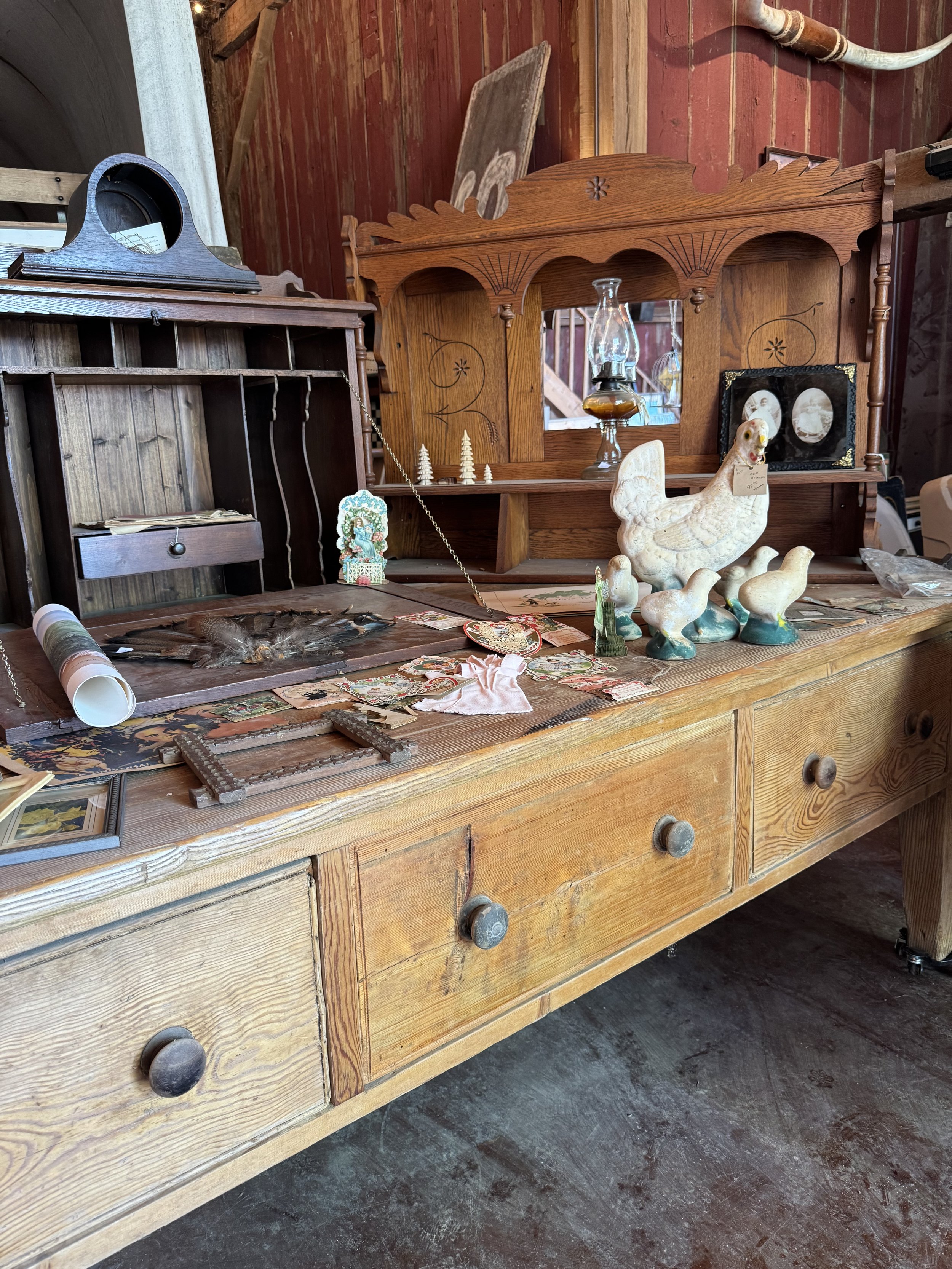 Vintage wooden dresser with various decorative items including figurines, picture frames, a lamp, and framed pictures. The background shows a wooden wall and additional antique furniture.
