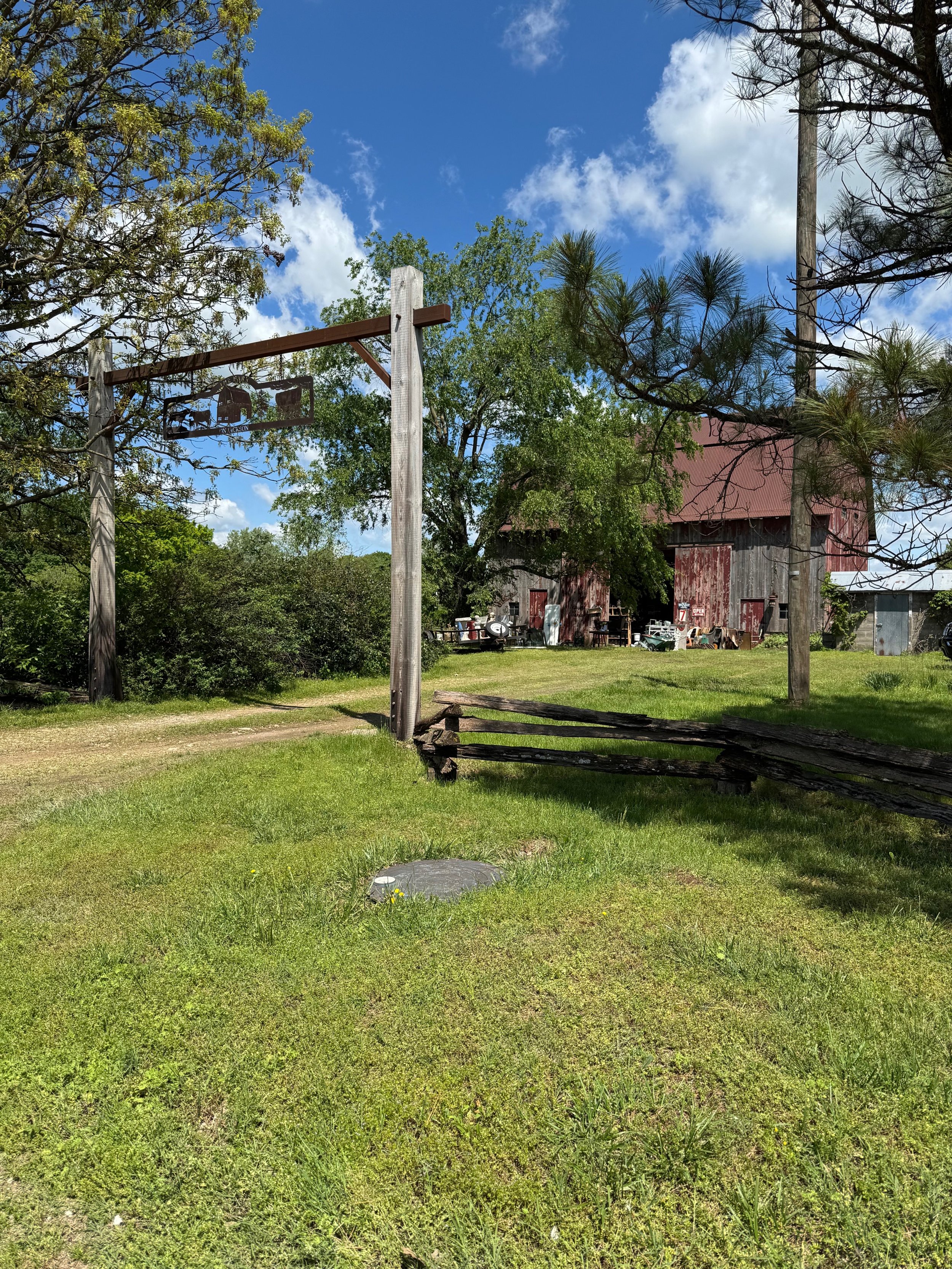 Scene of a grassy outdoor area with an old wooden barn in the background, a basketball hoop without a net, and trees around. The sky is blue with some clouds.