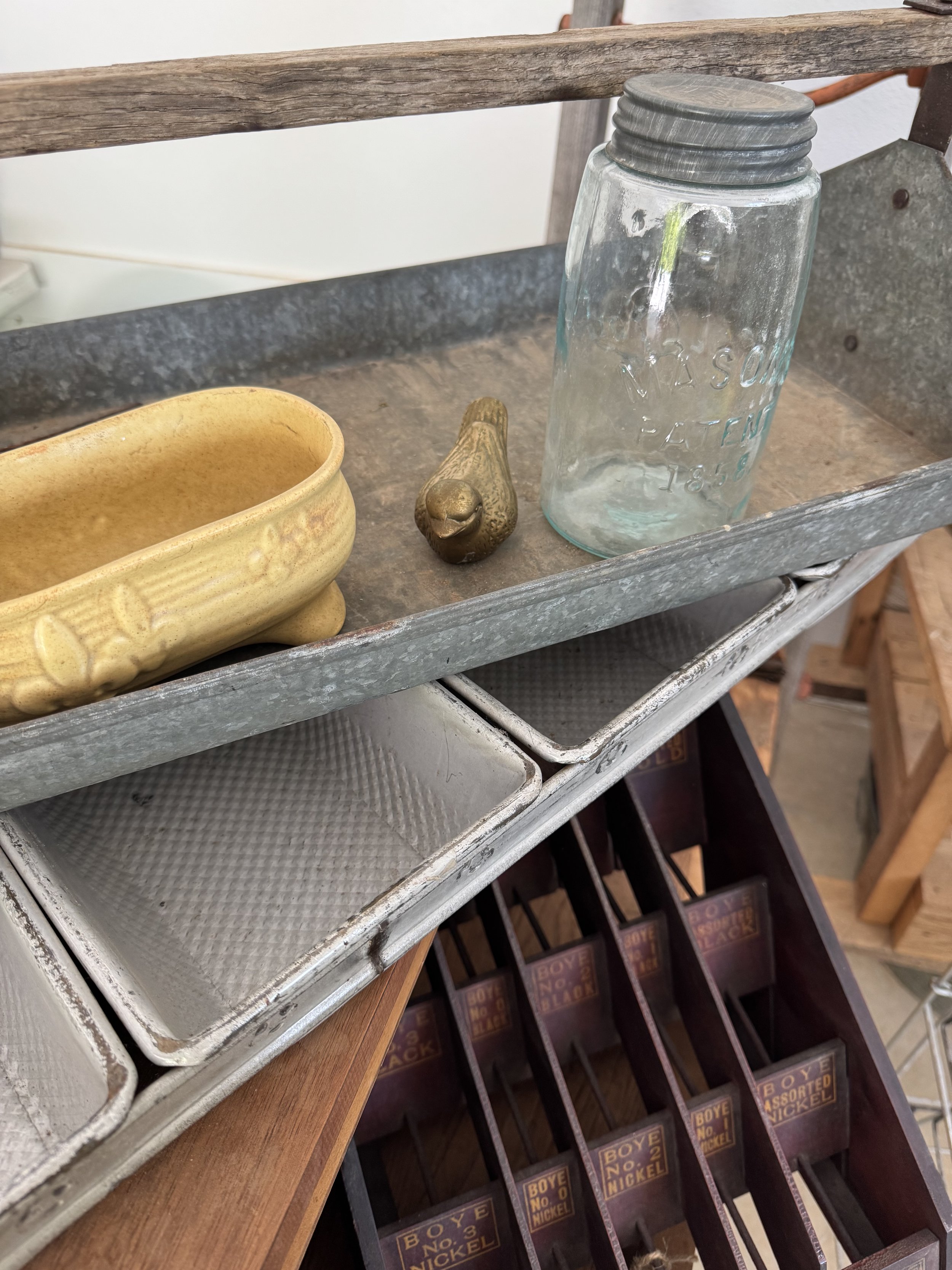 A rustic shelving unit holds a yellow ceramic dish, a small bronze bird figurine, and an old glass jar with a metal lid. Below, there are metal trays and compartments labeled for different types of nickel coins.