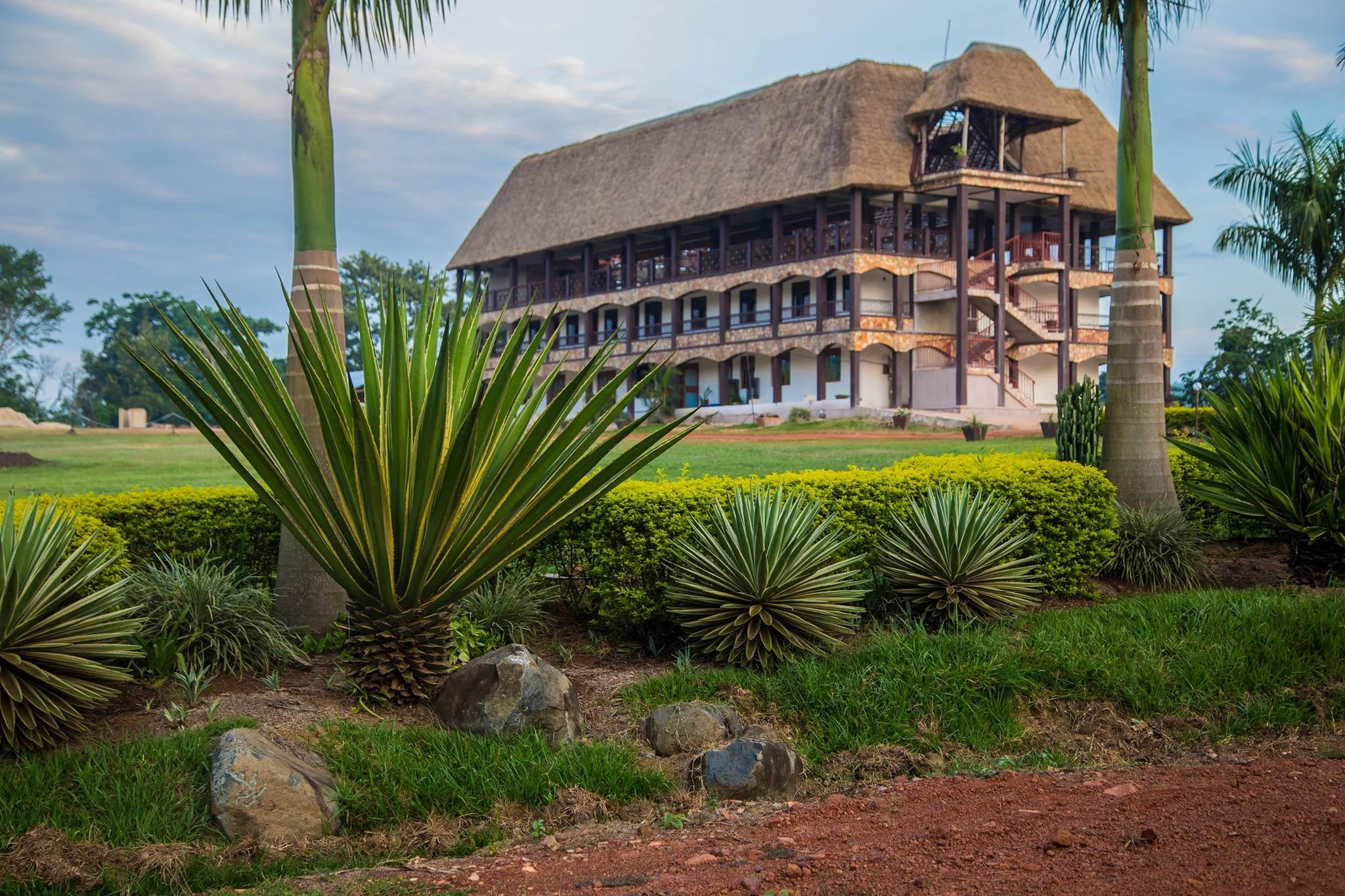 A large multi-story building with thatched roof and wooden balcony surrounded by lush greenery, including large spiky plants and palm trees.