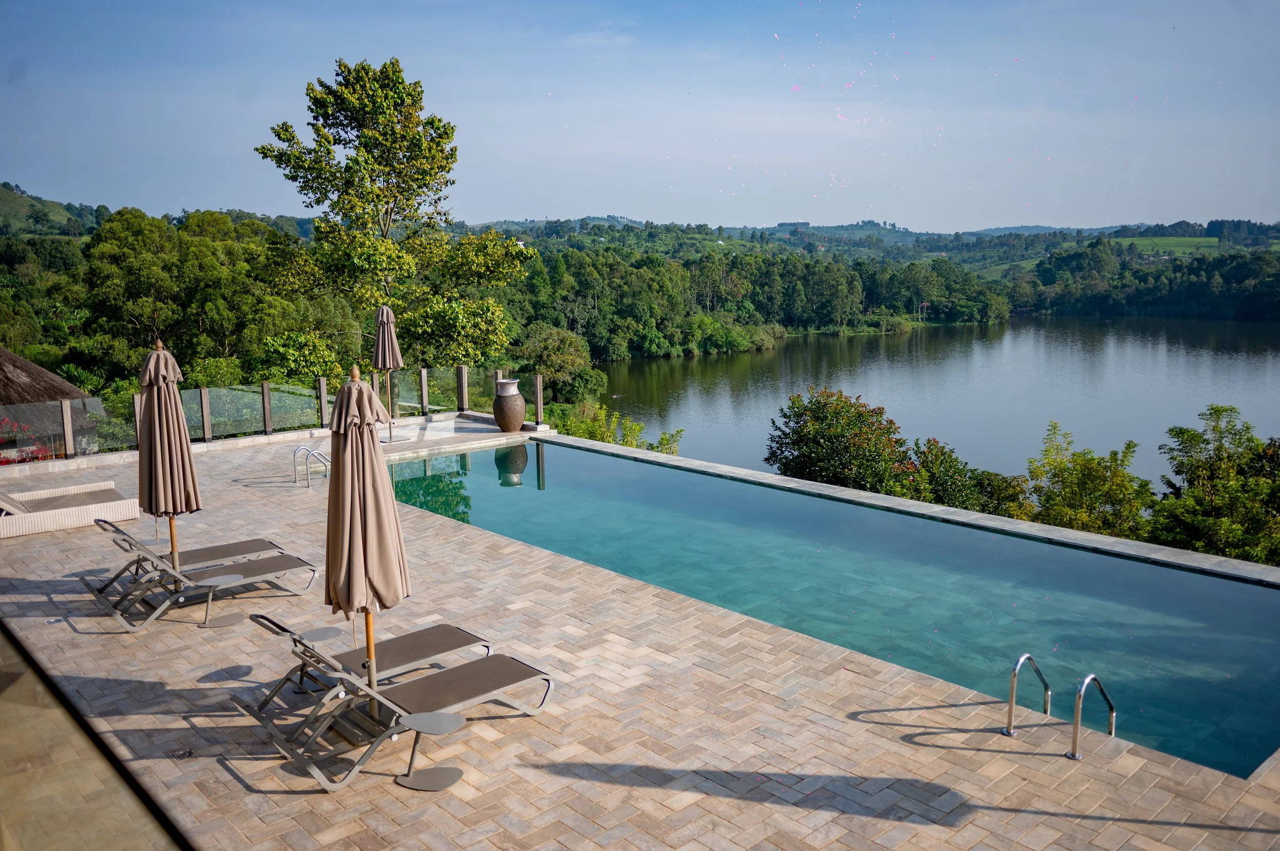 Infinity pool overlooking a river and lush green trees with rolling hills in the background, outdoor lounge chairs and umbrellas on a tiled pool deck.