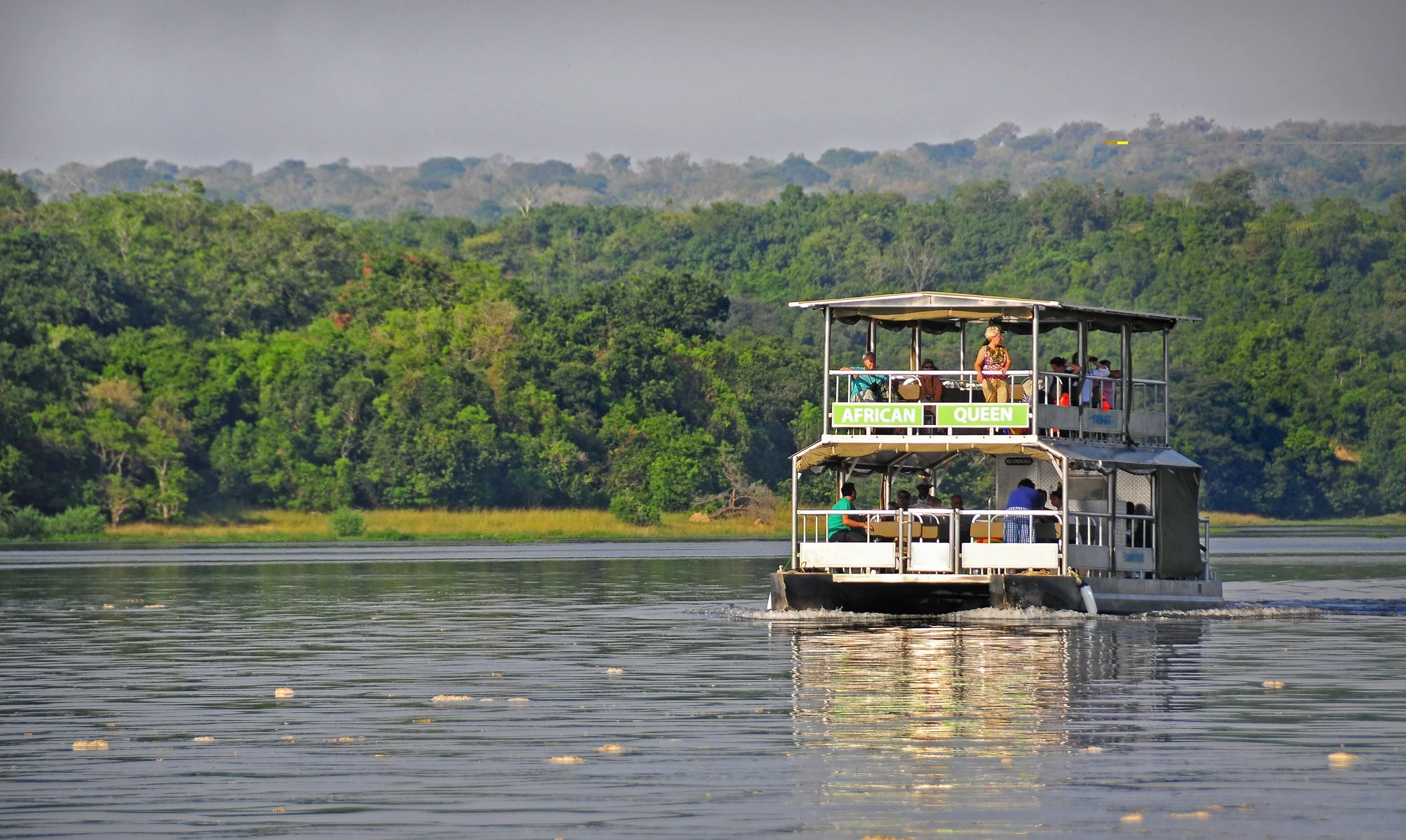 A two-story boat named 'African Queen' sailing on a river with lush green trees in the background. Passengers are seated on both decks.