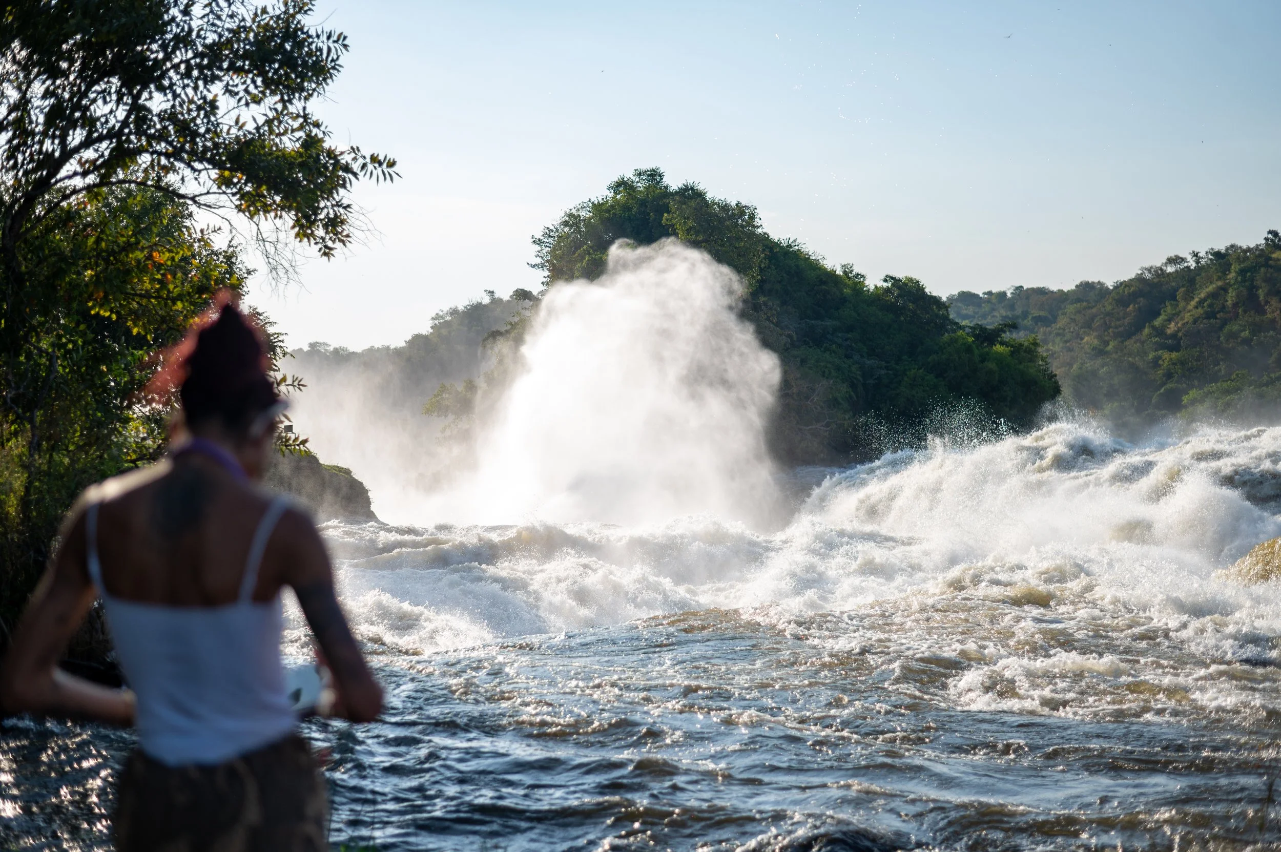 Person with a tattoo on their shoulder wearing a white tank top, standing by the river, watching the water waves and spray crashing against rocks with lush greenery in the background.