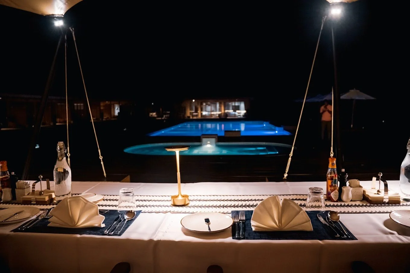 Elegant outdoor dinner table with folded napkins, glassware, and condiments at night, overlooking a lit swimming pool and hot tub.