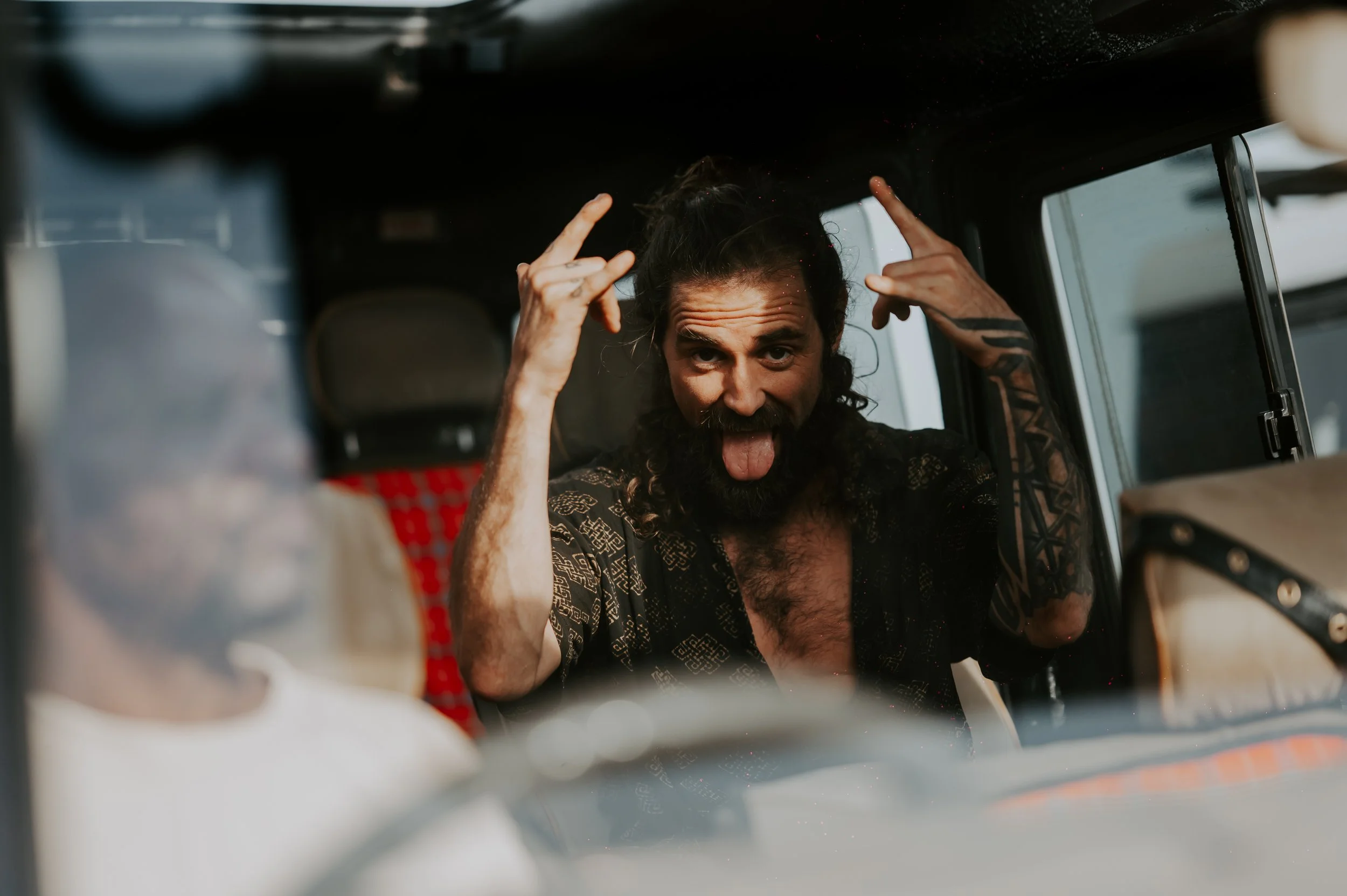 Man with long hair and beard making a rock and roll gesture in front seat of a vehicle, sticking out his tongue.