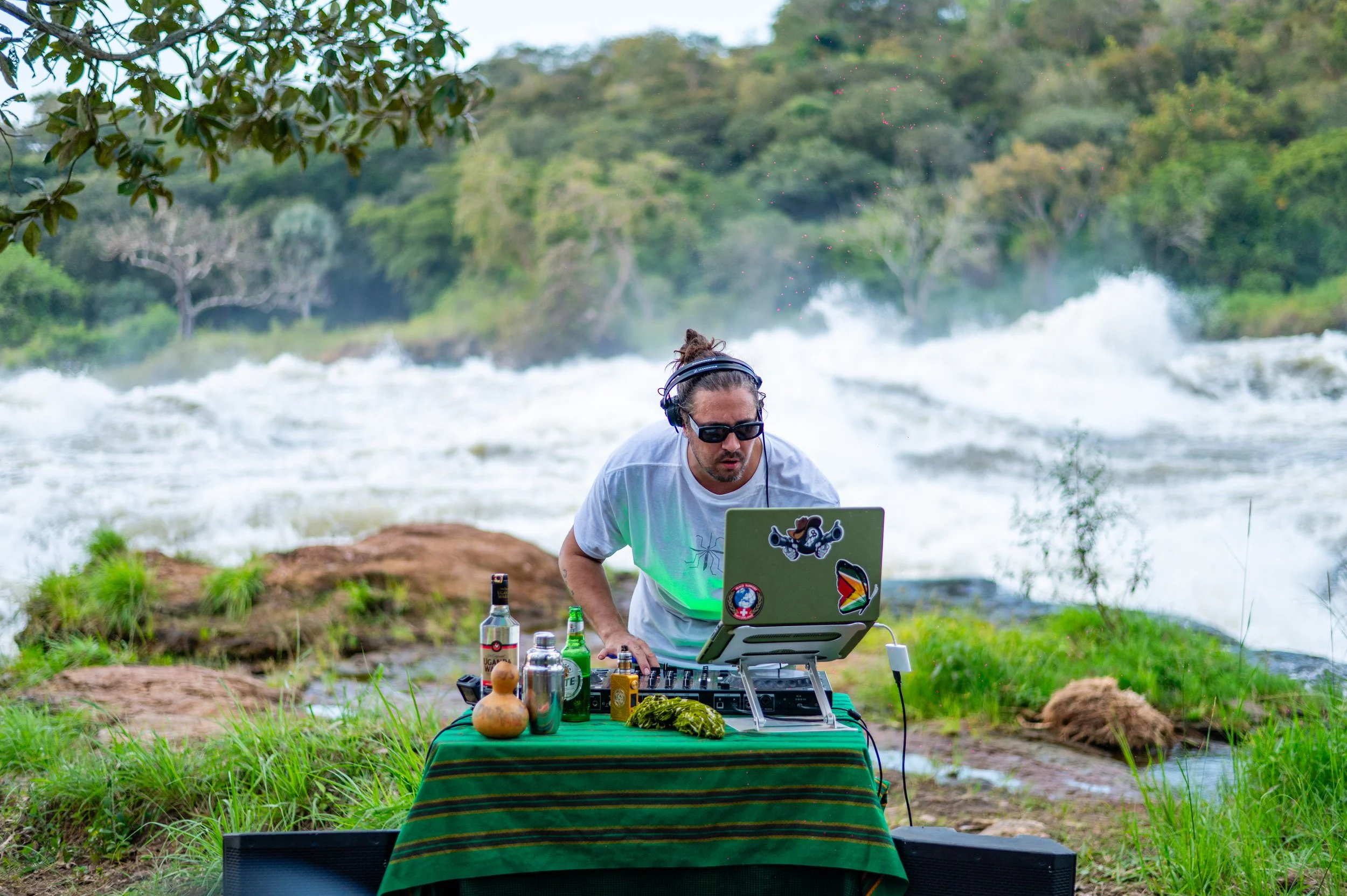 DJ wearing sunglasses and headphones, mixing music on a laptop set up on a green table outdoors near a river with rapids, surrounded by trees and grass.