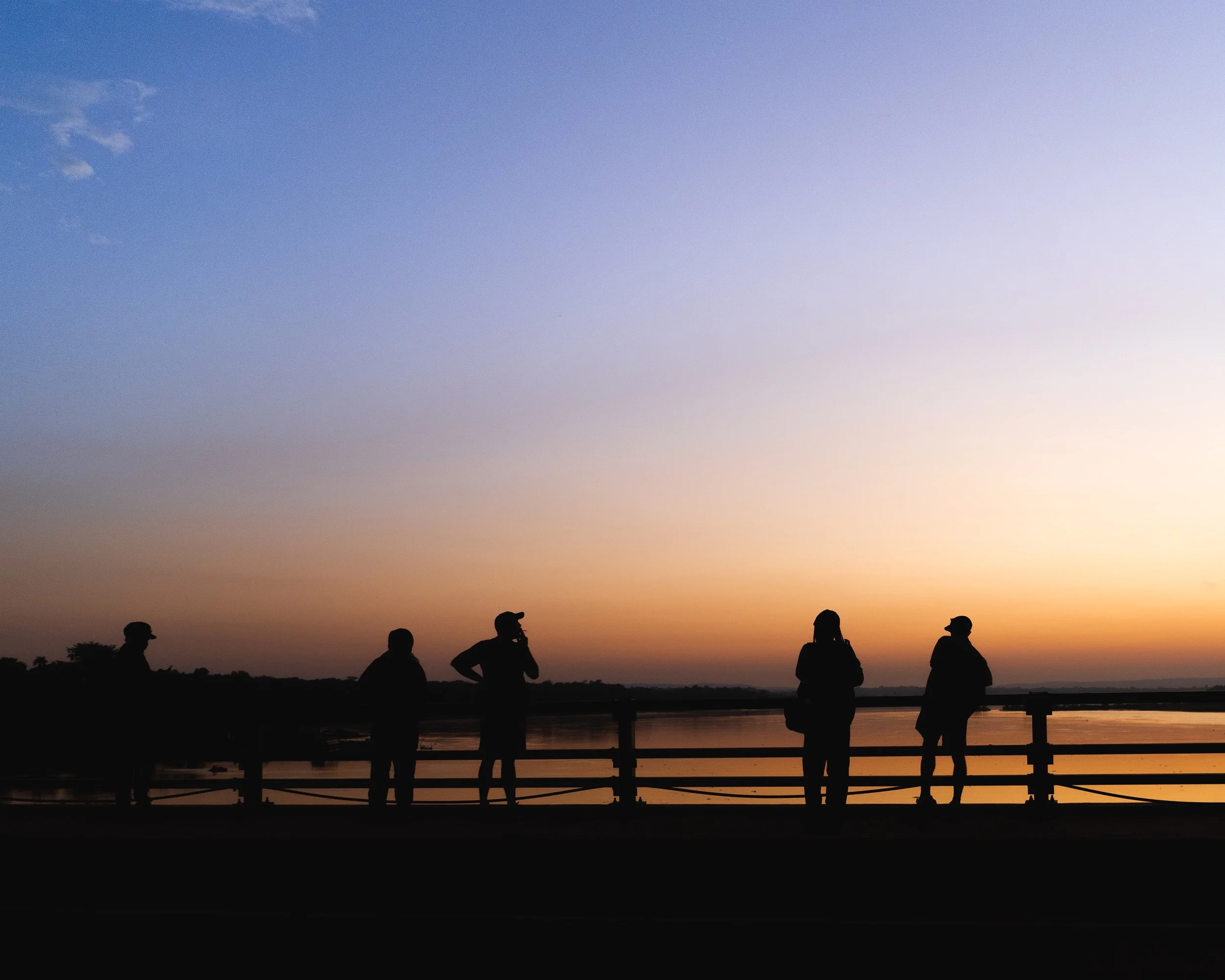 Silhouettes of five people standing by a railing near water during sunset, with a colorful sky in the background.