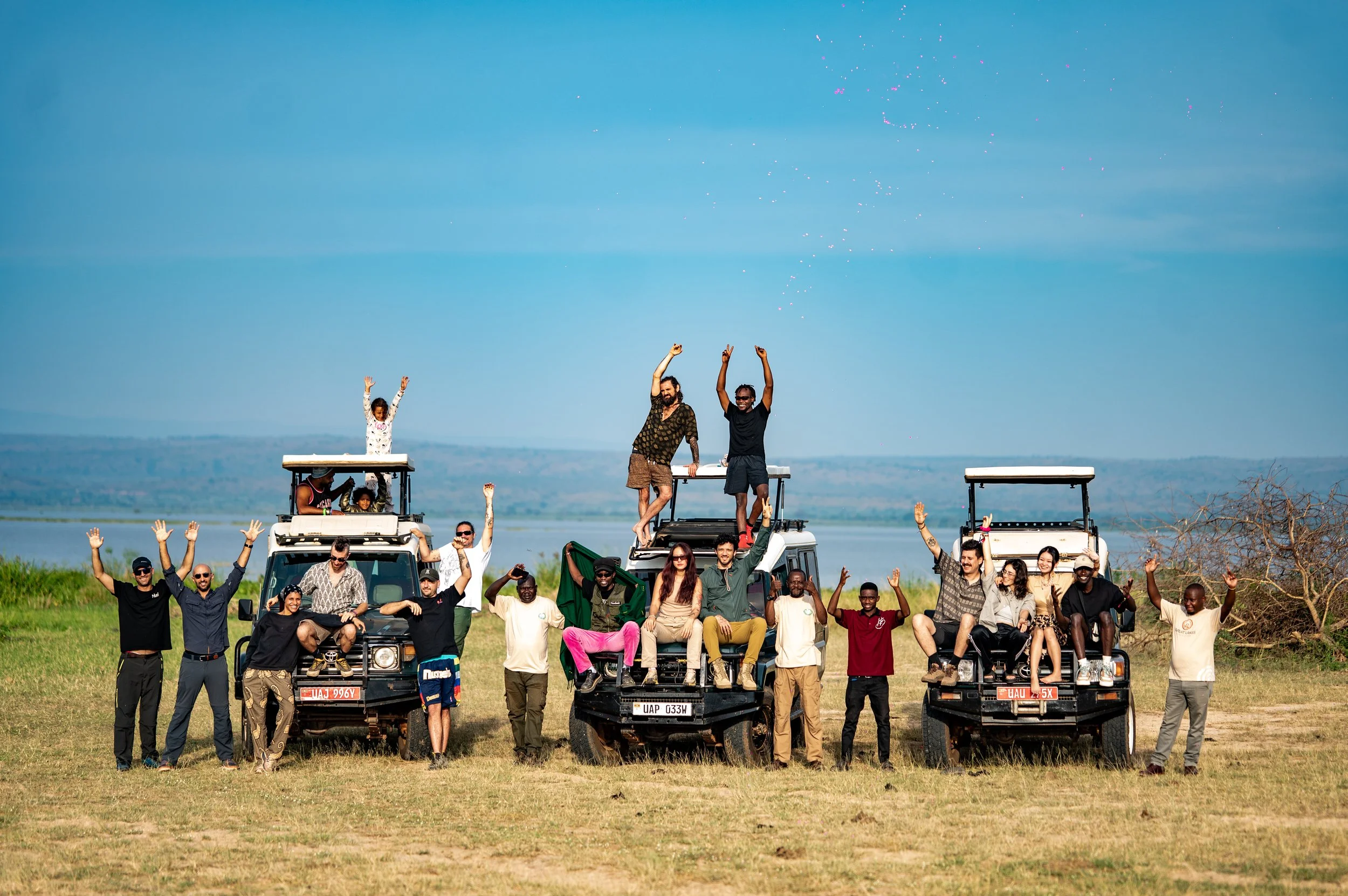 Group of diverse people celebrating outdoors with three off-road vehicles, some sitting and others standing, with arms raised and smiling, in an open field near a body of water.