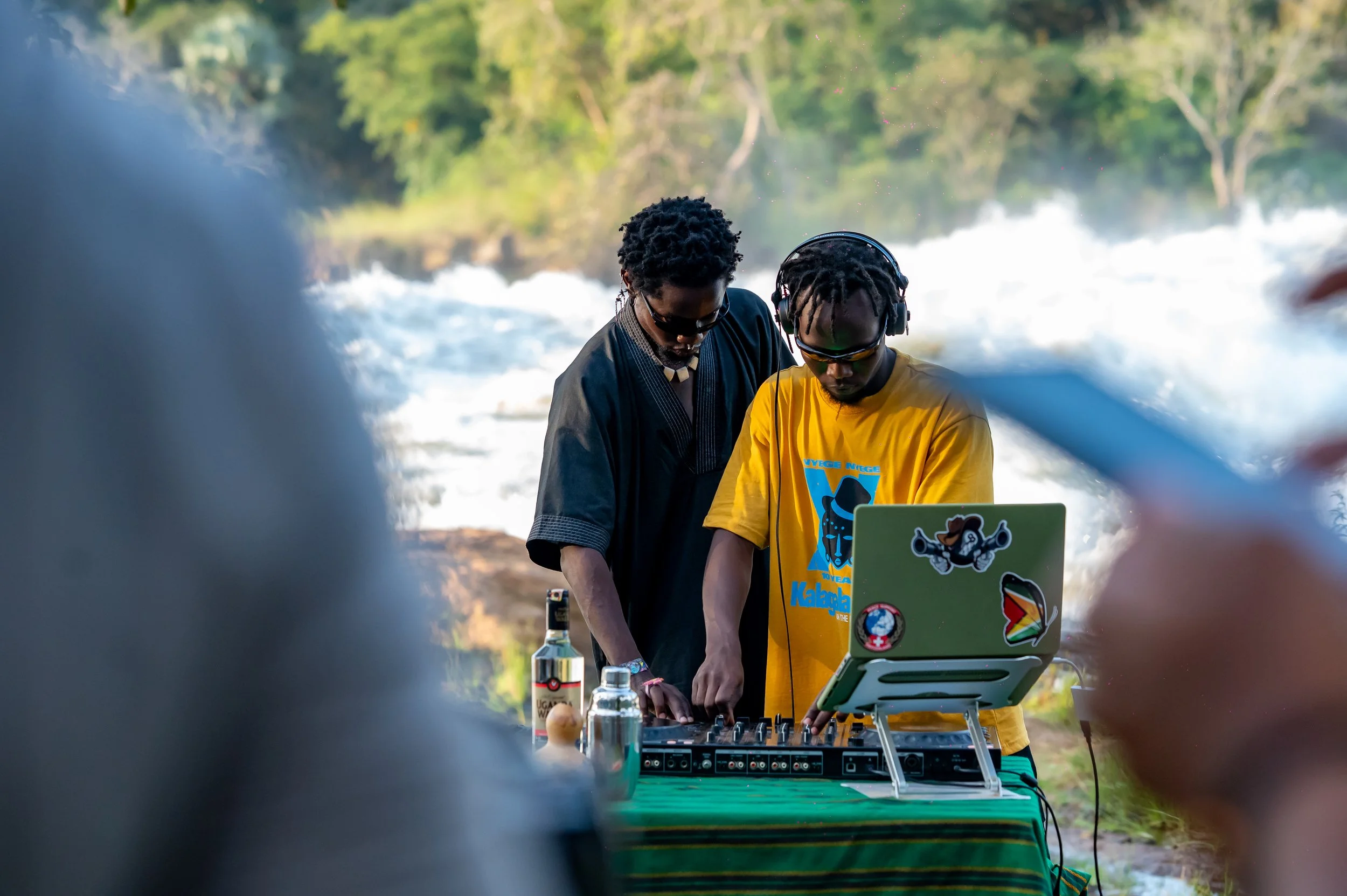 Two men are DJing outdoors near a river with a wooded background. One is wearing a black shirt and sunglasses; the other is wearing a yellow T-shirt, headphones, and sunglasses. There is DJ equipment, a laptop with stickers, and a bottle of liquor on