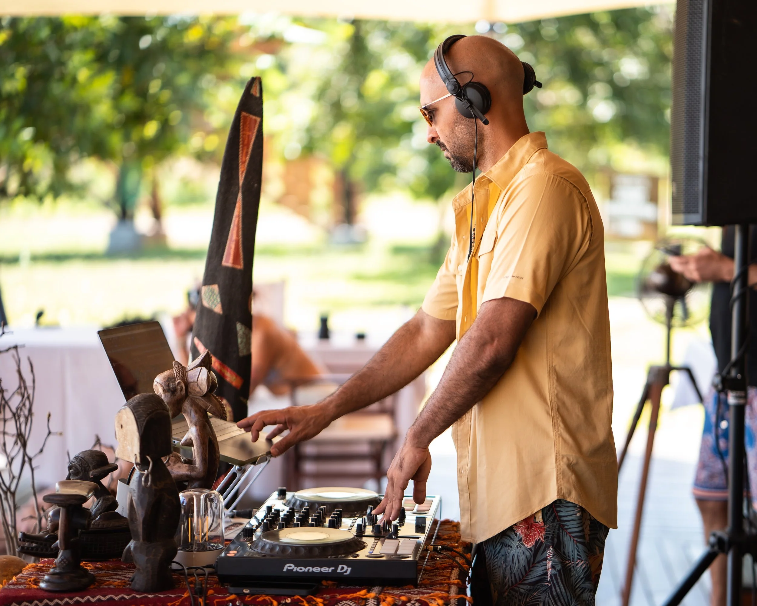 A DJ wearing headphones and sunglasses, dressed in a yellow shirt and patterned shorts, operating a DJ console outdoors with trees and people in the background.