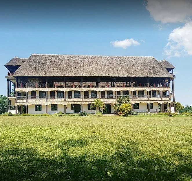 A large, multi-story building with a thatched roof, balconies, and tropical plants in front, set in a grassy field under a partly cloudy sky.