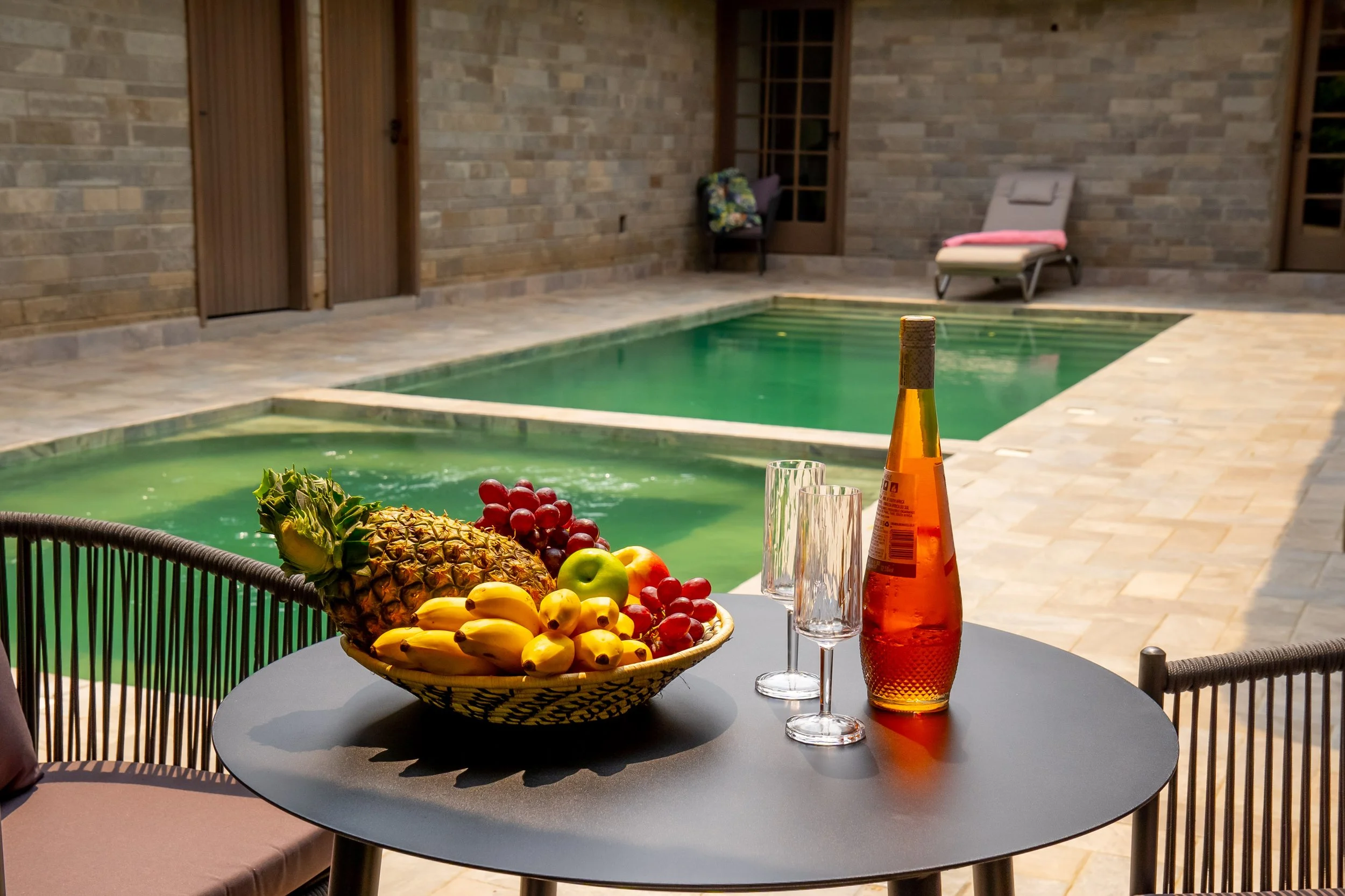 A table with a basket of assorted fruits, two glasses, and a bottle near a backyard pool with lounge chairs and a stone patio.