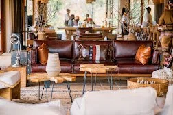 A modern living room with a brown leather sofa, a white vase on a side table, and a wooden coffee table. In the background, there are large windows and people sitting and standing in a social setting.