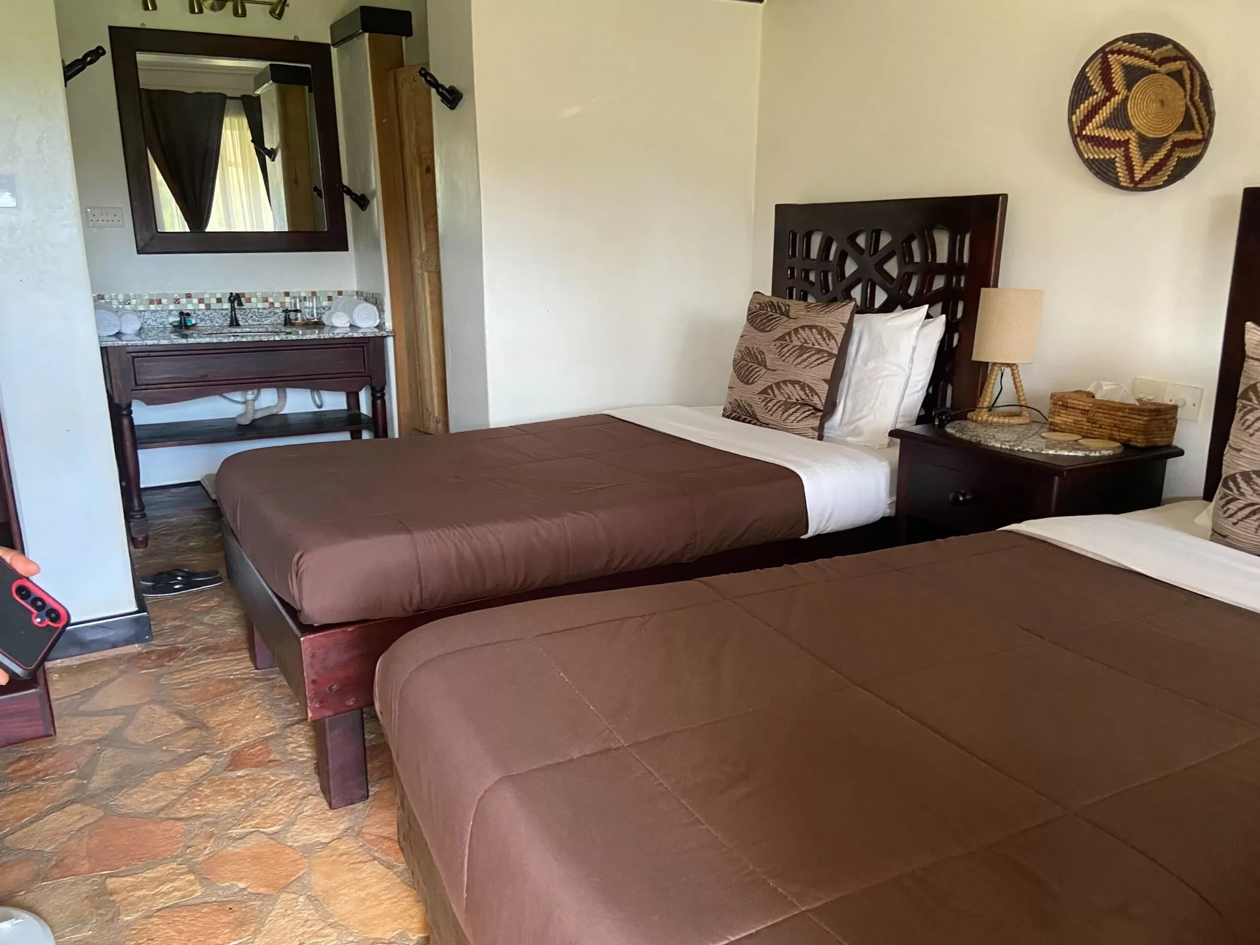 A hotel room with two beds with brown bedspreads, pillow with leaf print, a nightstand with a lamp and tissue box, a small mirror above a sink, and beige stone flooring.