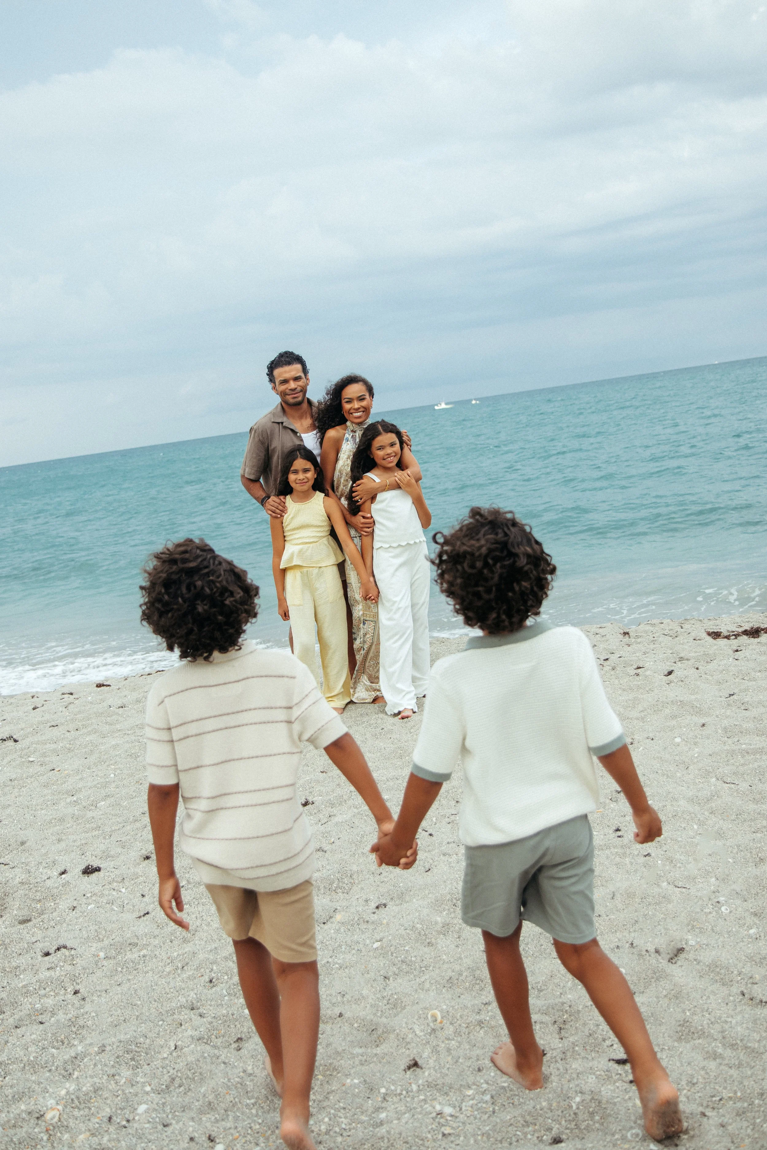 A group of children holding hands and walking on a sandy beach towards a beach family with an ocean view, cloudy sky, and boats in the sea during an Outdoor family Beach Photography Session