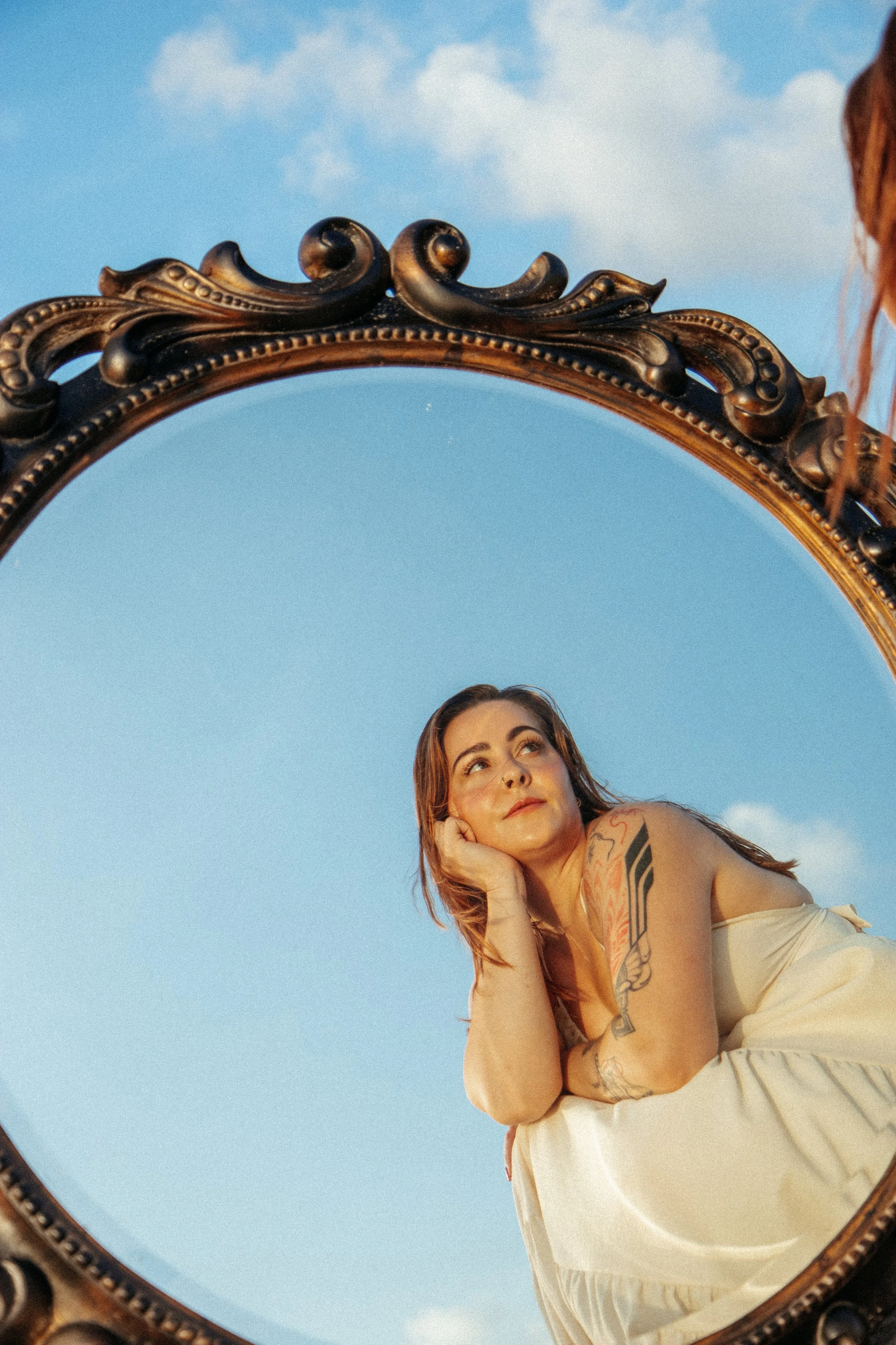 A woman with red hair and tattoos resting her chin on her hand in front of an antique mirror, with the sky and clouds reflected in the mirror.