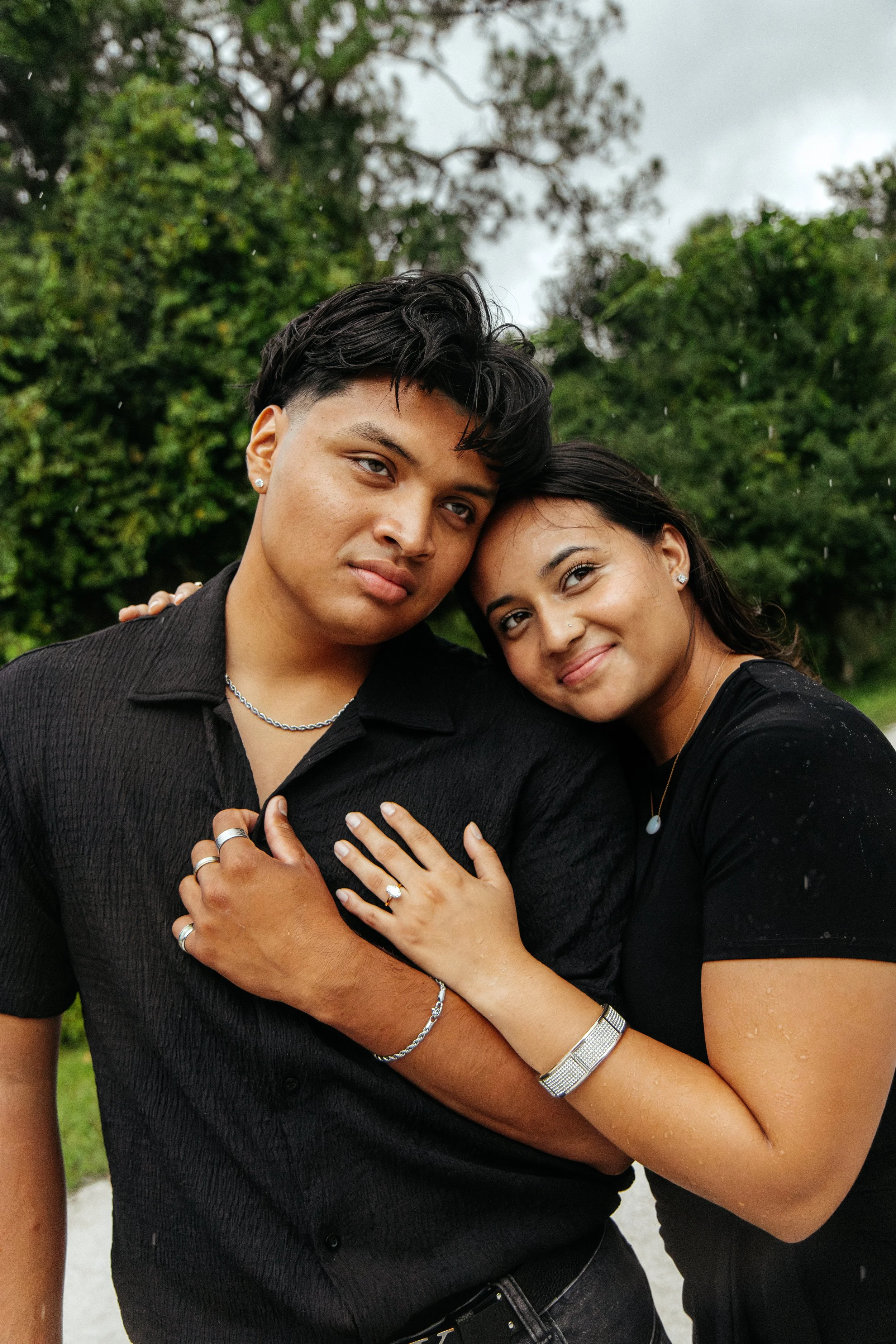 A young man and woman are close together outdoors, with the woman resting her head on the man's shoulder, both smiling gently, wearing black clothing with greenery in the background.