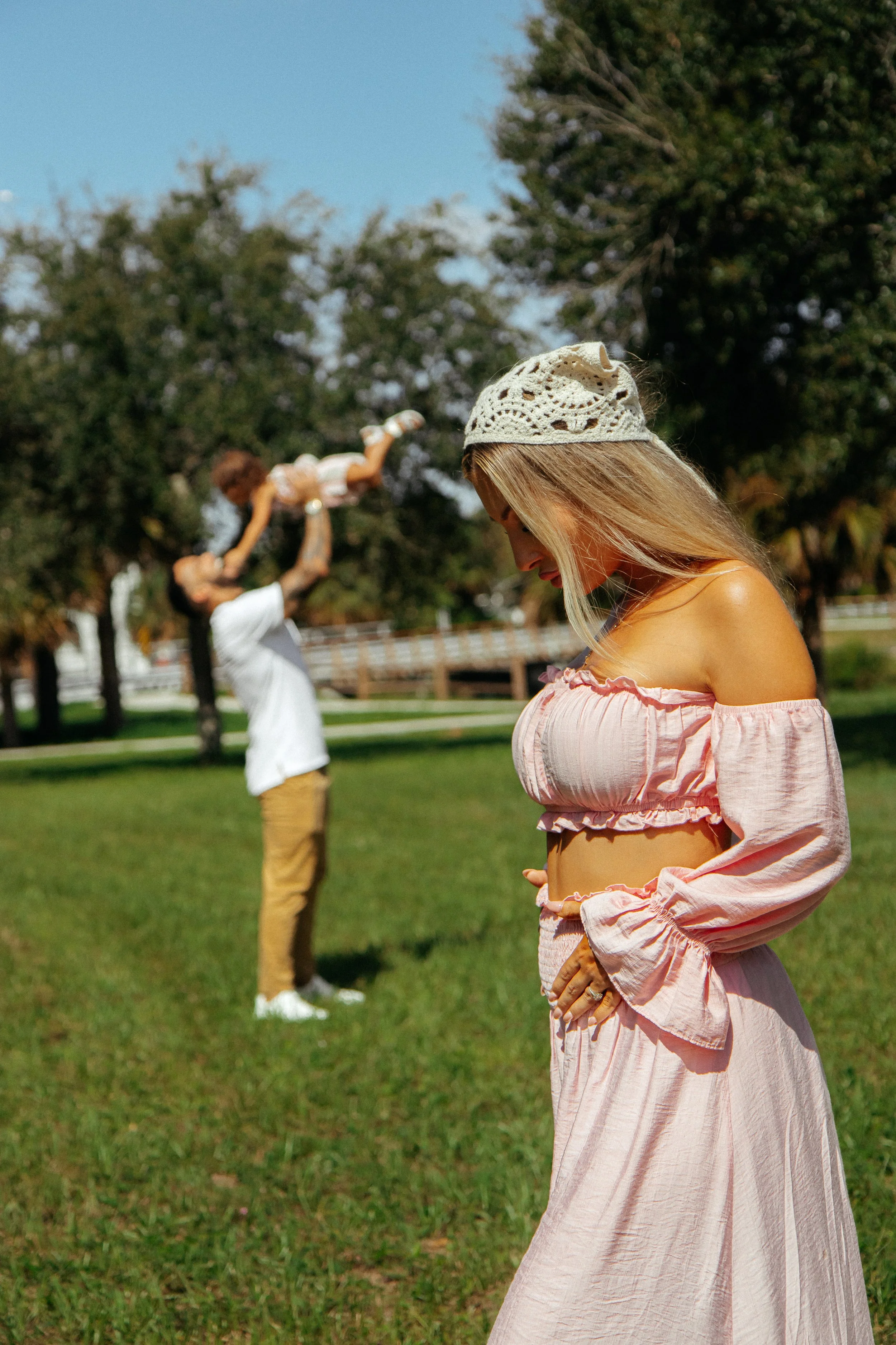 A woman wearing a pink off-shoulder dress and a white lace hat, standing outdoors on grass with her head bowed. In the background, a person is playing with a dog, lifting it into the air, with trees and a fence behind them.