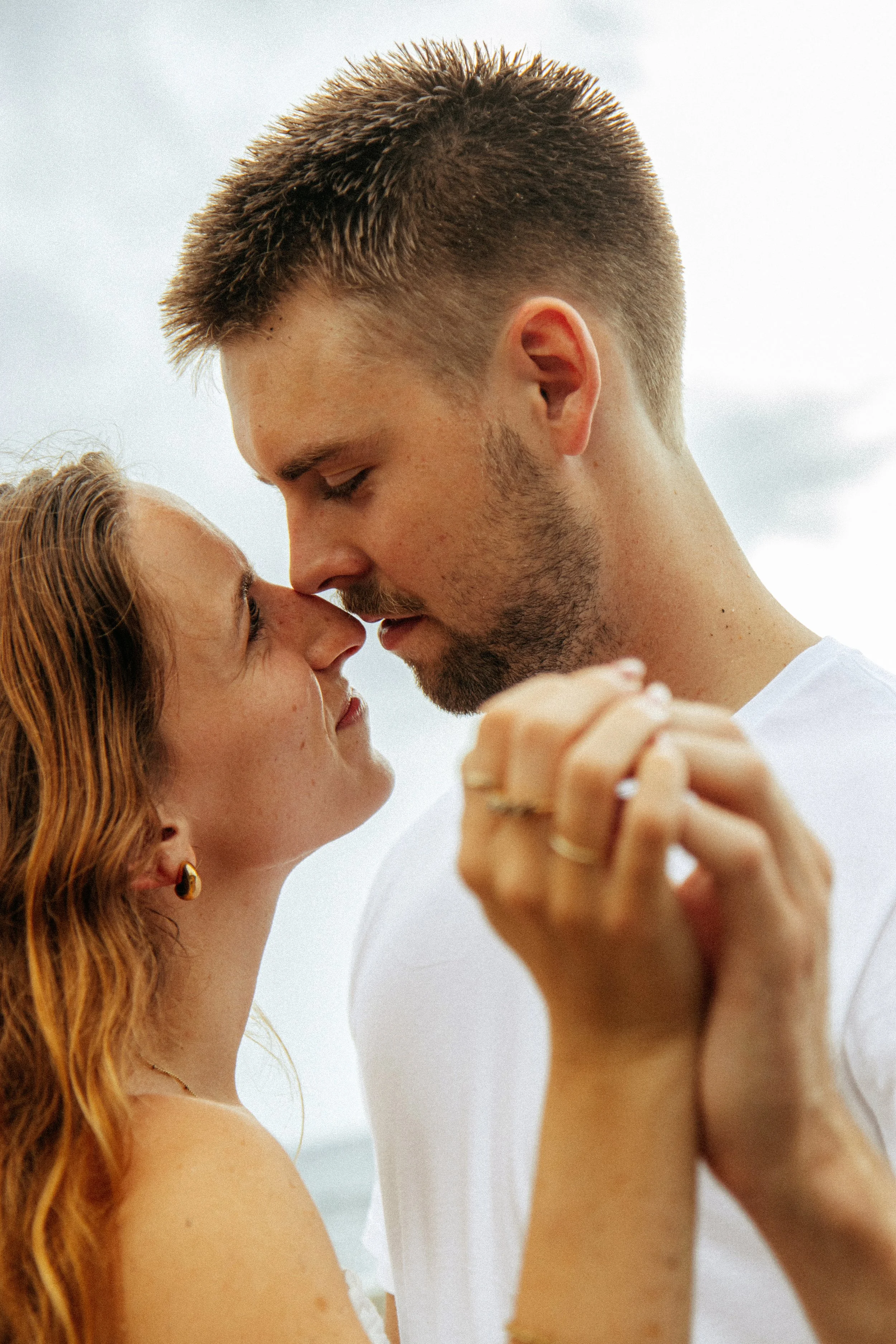 A close-up of a young couple with their foreheads touching and eyes closed, holding hands, outdoors against a cloudy sky durning a Romantic Beach Couples Photography Session