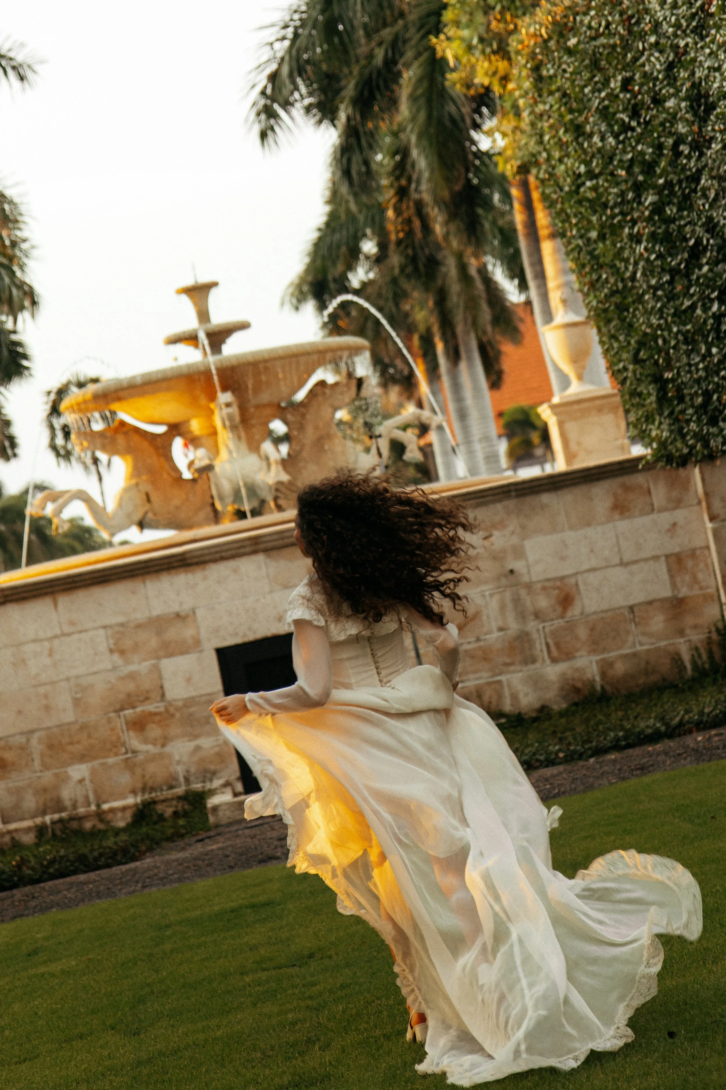 A woman in a flowing white gown is walking in front of a stone fountain surrounded by palm trees and bushes at sunset.