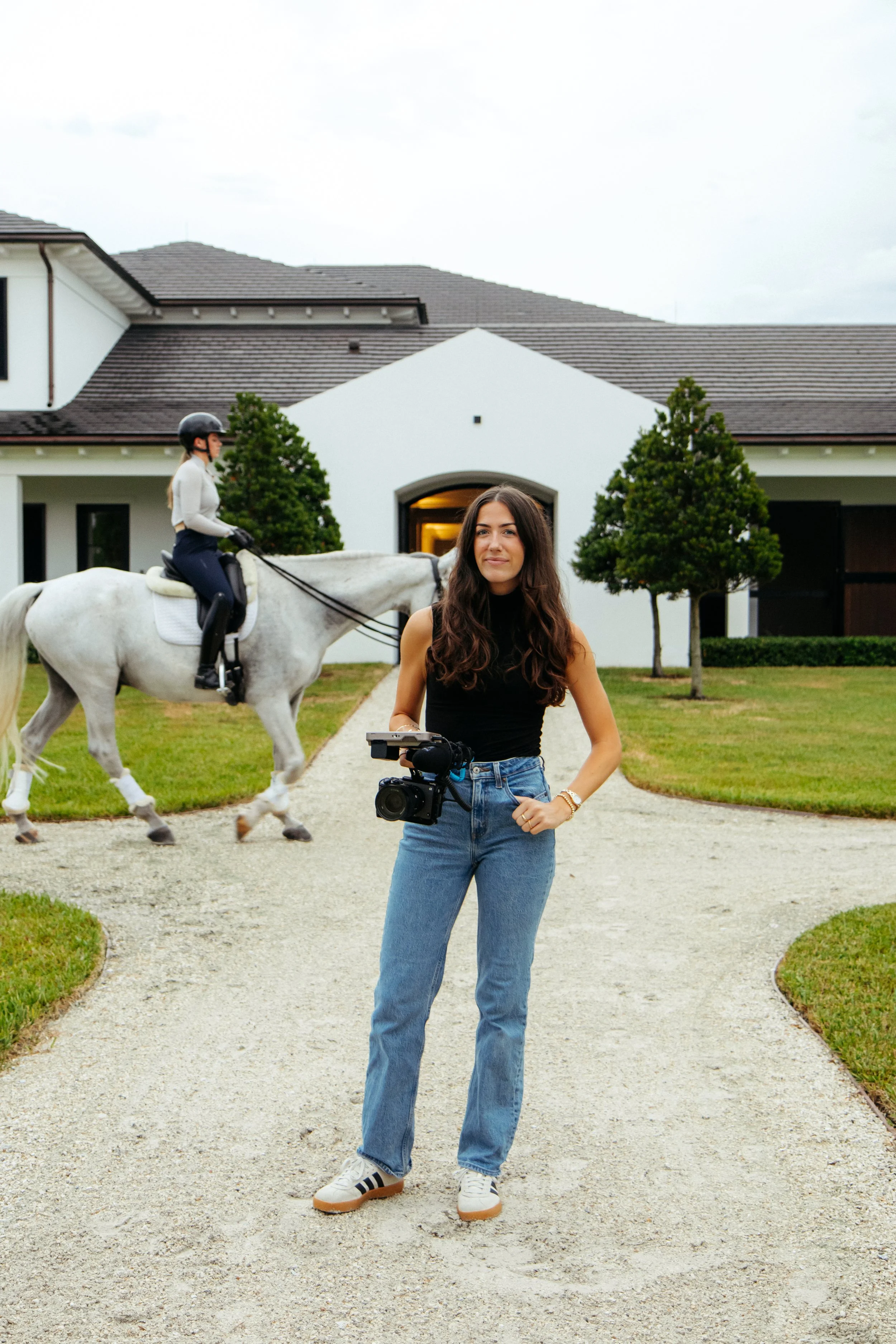 A woman with long brown hair wearing a black sleeveless top and blue jeans standing on a gravel pathway in front of a large white house with dark roofs. Behind her, a person riding a white horse, wearing riding gear and a helmet, is visible. There ar