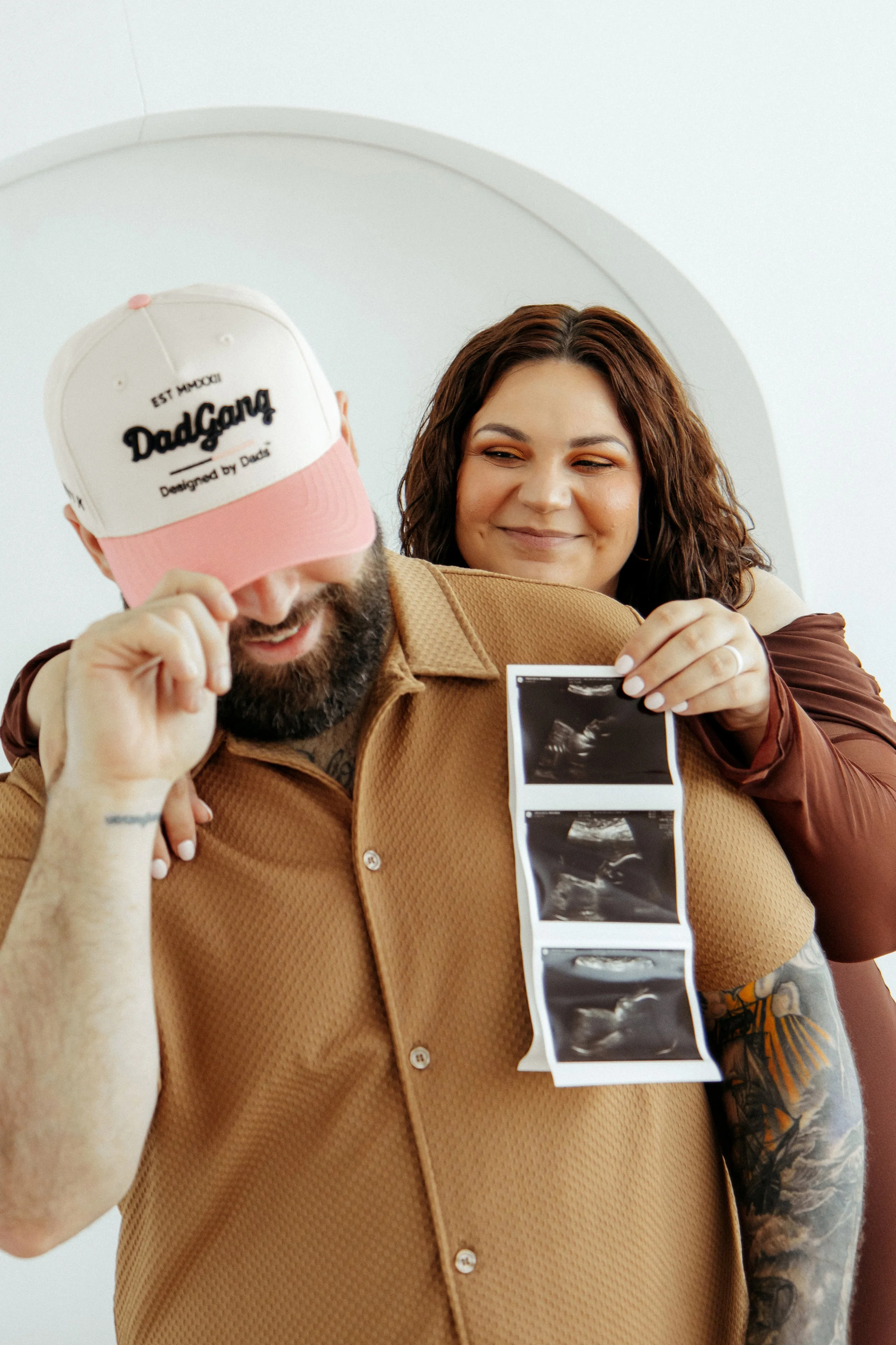 A man and woman celebrating with ultrasound photos, the man is holding a cap with 'DadGang' written on it, and the woman is smiling during an Indoor Maternity Photography Session