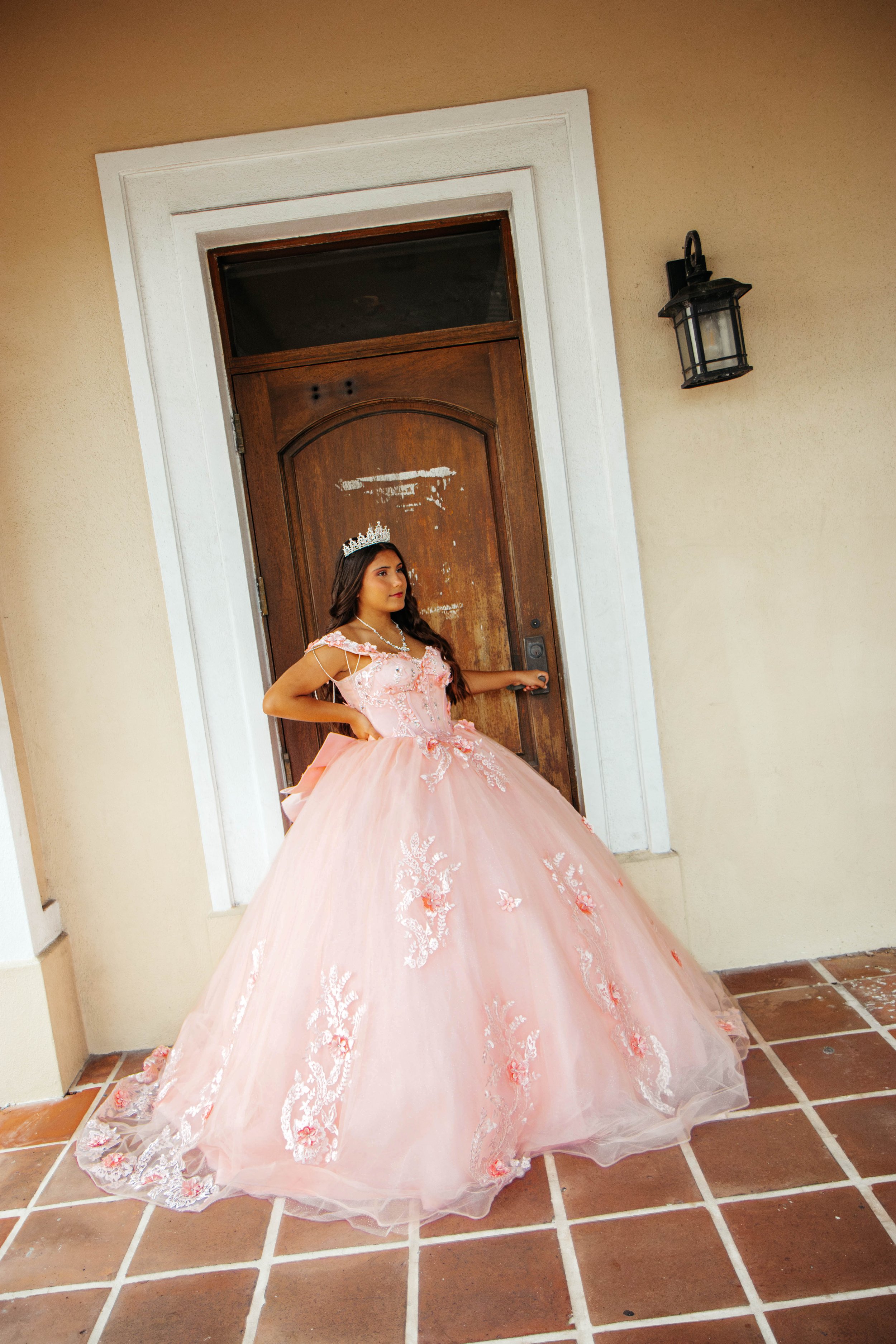A young woman in an elegant, pink, floral-embellished ball gown with a tiara standing outside a house near a wooden door with a black lantern light fixture on the wall.
