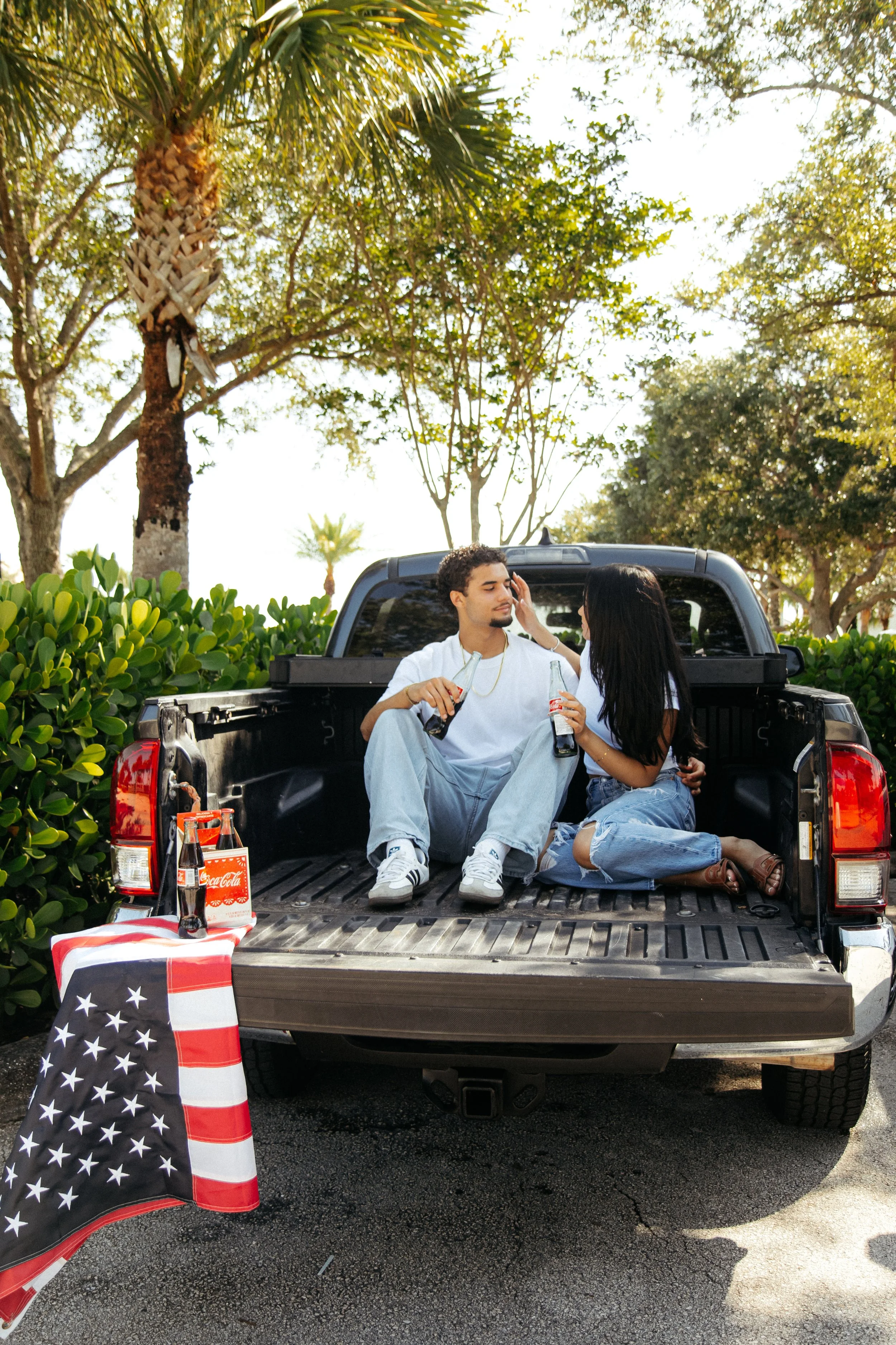 A young man and woman sitting in the bed of a pickup truck, drinking soda bottles and touching each other's faces in a park-like setting with trees and greenery. There is an American flag and soda bottles on a small table nearby.